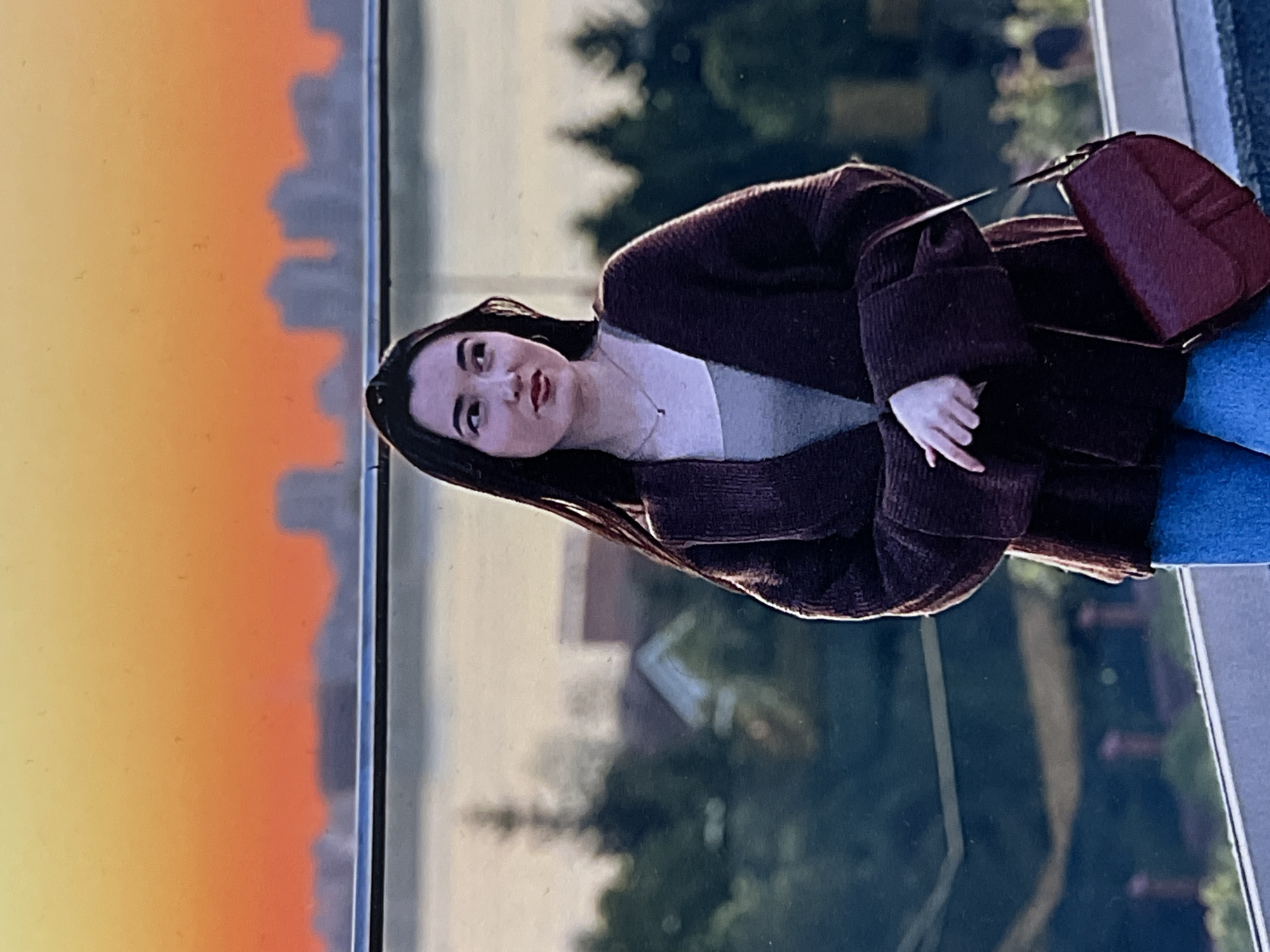 A young woman is depicted in a portrait, positioned in what appears to be an urban setting during sunset. She has long, dark hair, and is wearing a dark brown ribbed cardigan over a light-colored top, paired with blue jeans. A reddish-brown shoulder bag is visible on her left side, and she wears a delicate silver necklace with a small pendant. Her expression is neutral, with slightly parted lips showing a hint of red lipstick, as she looks slightly to her right.
The woman is standing in front of a large window or glass barrier. Through the glass, on her left, a striking sunset paints the sky in shades of yellow, orange, and red. Silhouetted against this colorful backdrop are numerous city buildings, suggesting a dense urban landscape, characteristic of Kazan, Russia. To her right, beyond the glass, blurred green trees and architectural forms are visible. The scene implies an elevated viewpoint, possibly from a building or observation deck overlooking the city. The time of day is clearly late afternoon or early evening, marked by the dramatic sunset. The woman appears to be posing for the camera. There is no visible text in the image.