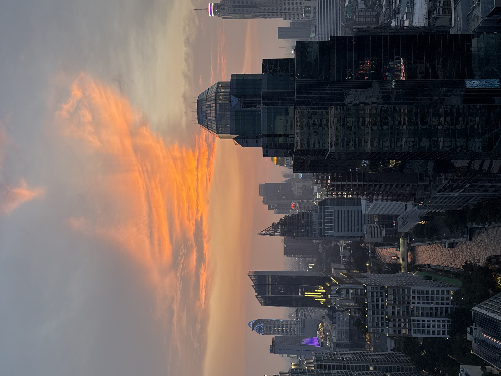 The image shows a cityscape at dusk, likely Bangkok, Thailand, given the architectural styles of the buildings. A tall, glass-faceted skyscraper dominates the center, flanked by other high-rise structures. The sky displays streaks of orange and grey clouds, suggesting either sunrise or sunset. A river or canal meanders through the lower part of the city, reflecting the city lights. Illumination from buildings is visible, indicating the transition from day to night. There are no discernible people in the scene.