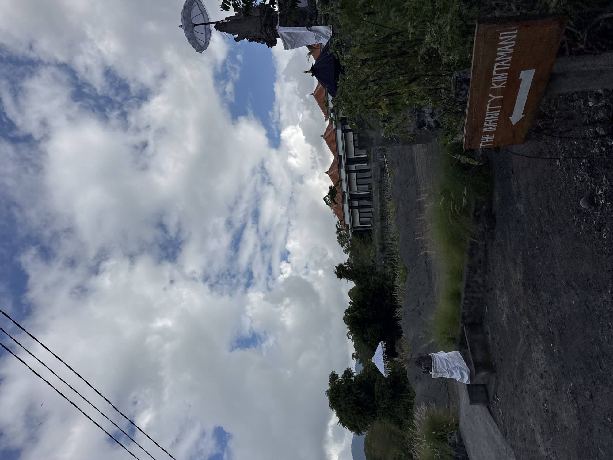A low-angle outdoor shot captures a cloudy day in Kintamani, Indonesia. In the foreground, a weathered wooden sign with white lettering reads "THE INFINITY KINTAMANI" and features a white arrow pointing to the left. The sign is mounted on a wooden post. To the left of the sign, a pathway leads towards a small stone structure topped with a statue draped in white fabric, possibly an offering. Beyond this, a wide area of dark, loose soil or ash is visible, with tufts of dry grass growing sporadically. In the midground, a row of modern buildings with orange tiled roofs stands against a backdrop of lush green trees and distant mountains. Several white and black umbrellas, characteristic of Balinese cultural elements, are placed near the buildings and on a stone structure to the right. Power lines stretch diagonally across the upper left quadrant of the sky. The overall impression is of a tranquil, scenic location, likely a resort or cultural site, under a dynamic sky.