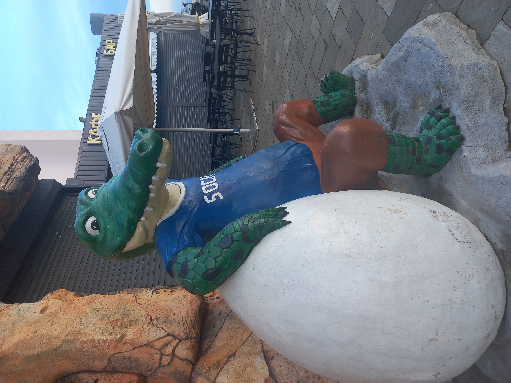 The image shows a statue of a green crocodile wearing a blue "SOCHI" jersey, lying on a large white egg. The statue is positioned in front of a building with a sign that reads "КАФЕ БАР" (Cafe Bar). Tables and chairs are visible behind the building. The statue appears to be part of a tourist attraction in the city of Sochi, Russia.