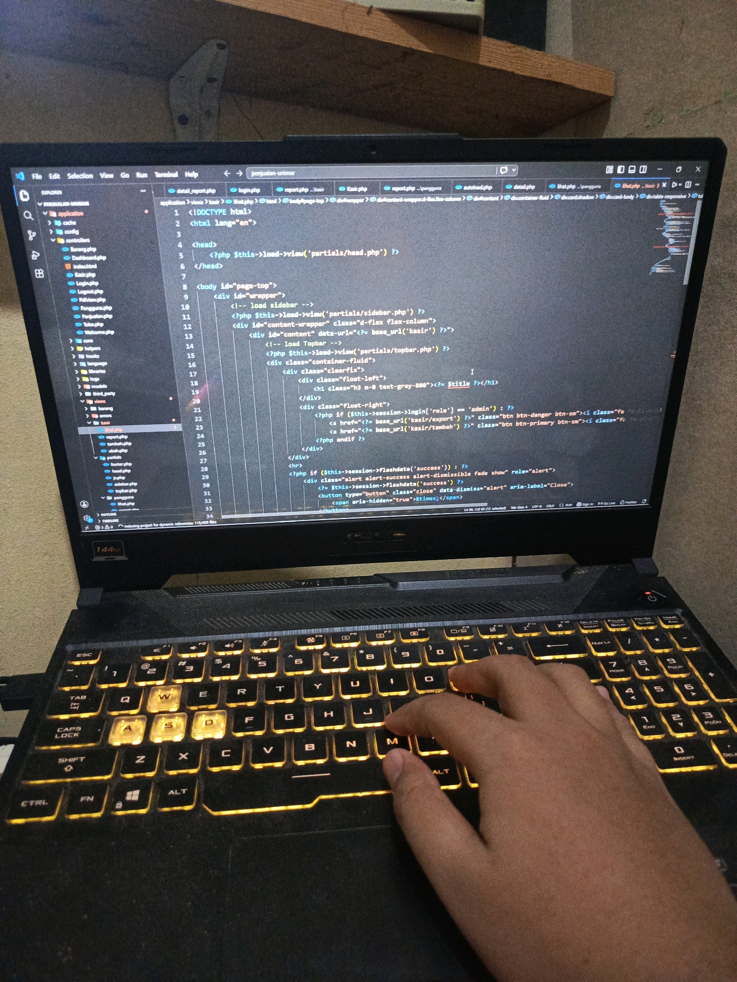 The image shows a close-up of a laptop with an illuminated keyboard and a hand resting on it. The laptop screen displays code, specifically PHP and HTML, within a code editor. The left side of the screen shows a file explorer with a directory structure. The background is out of focus, but appears to be a wooden surface. This scene is set in Balaraja, Indonesia, likely within a home or office environment. The overall impression is one of focused work or study related to web development.