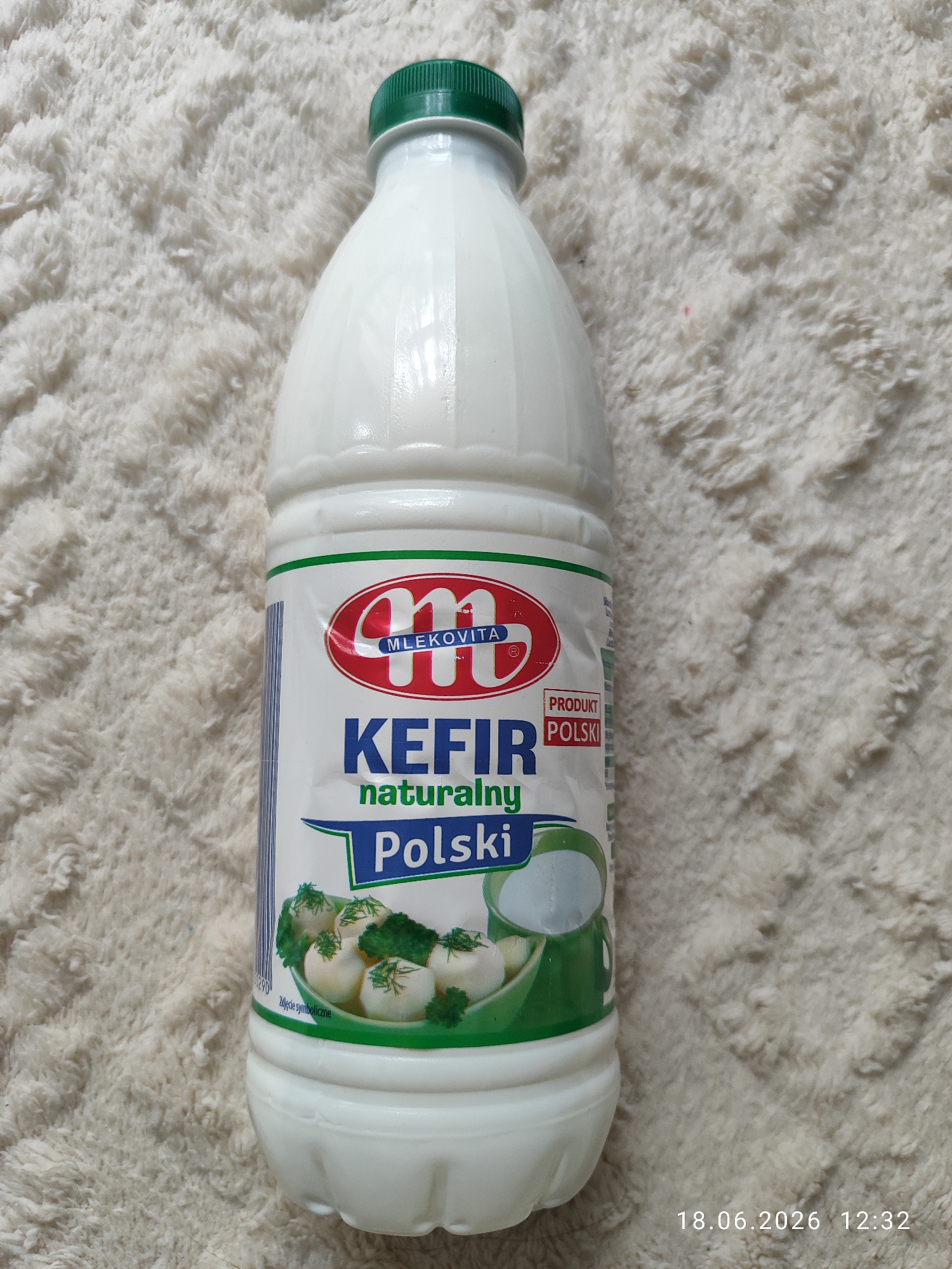 A white plastic bottle of Mlekovita Kefir Naturalny Polski is positioned upright on a white fluffy blanket. The bottle has a green cap. The label on the bottle is predominantly white with red and blue accents. At the top of the label, there is a red oval logo with the letter "M" in white. Below this, the text "MLEKOVITA" is visible.
The central part of the label features the word "KEFIR" in large blue letters, followed by "naturalny" in smaller green letters. Underneath "naturalny," the word "Polski" is written in large blue letters within a green banner. To the right of "KEFIR," a small red rectangle contains the text "PRODUKT POLSKI" in white letters.
At the bottom of the label, there is an illustration of a bowl containing small white balls, likely cheese, garnished with dill and broccoli. A green pitcher is also depicted. The text "Zdjęcie symboliczne" (Symbolic photo) is visible near the bottom edge of the illustration.
The bottom right corner of the image shows a timestamp: "18.06.2026 12:32".
The lighting appears to be artificial, and the scene is likely indoors. The overall impression is of a product shot, focusing on the kefir bottle. There are no people visible.