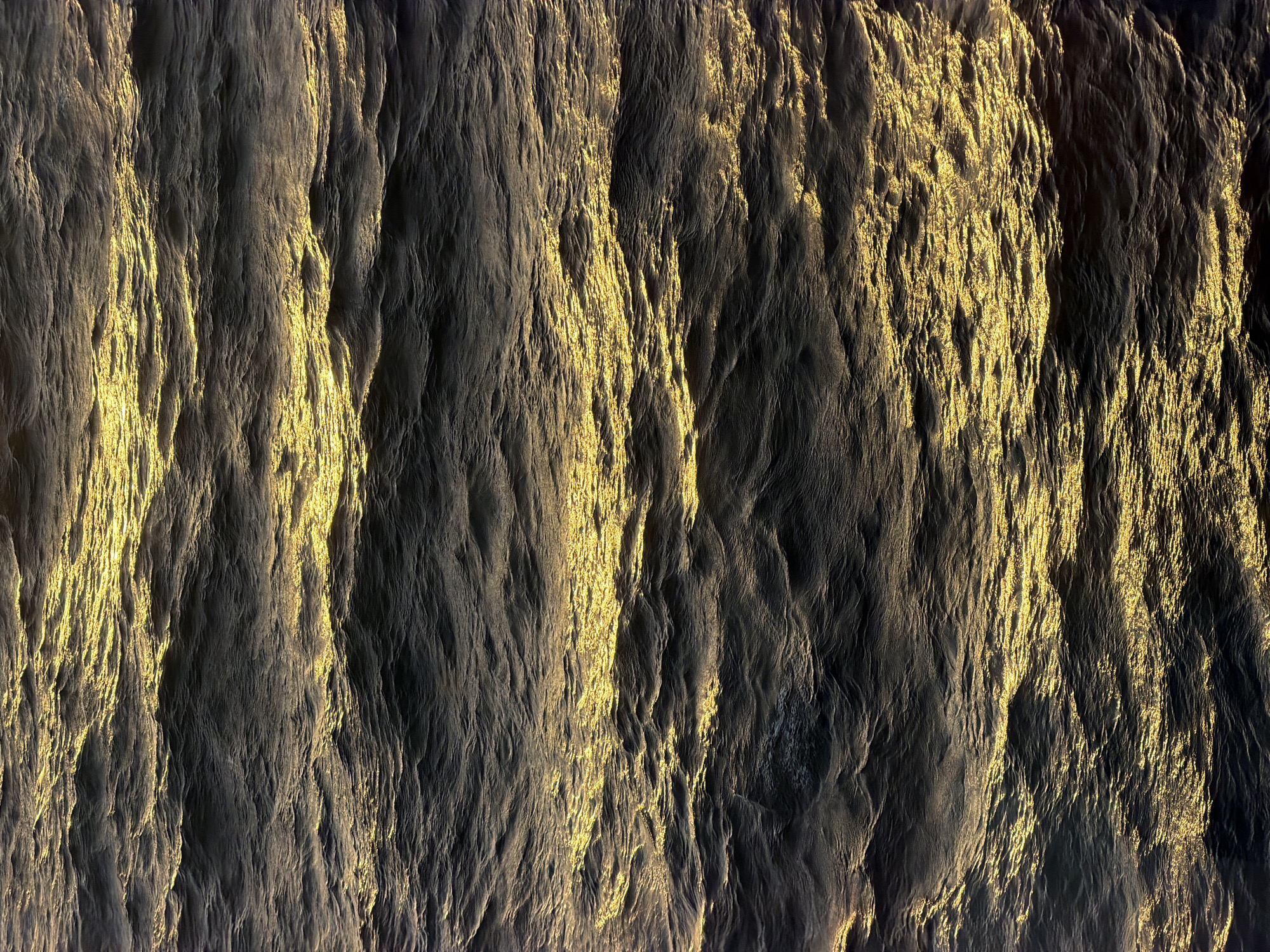 The image shows a textured surface, possibly of sediment or rock, with a prominent pattern of vertical striations. The color palette ranges from dark grays to light golds, suggesting that light is playing a significant role in defining the texture and highlighting ridges or grooves. The overall effect is reminiscent of a geological formation or a close-up view of a weathered material. There are no visible people, subjects, objects, actions, or interactions. The environment is limited to the textured surface itself. The location is Unknown, Unknown.
