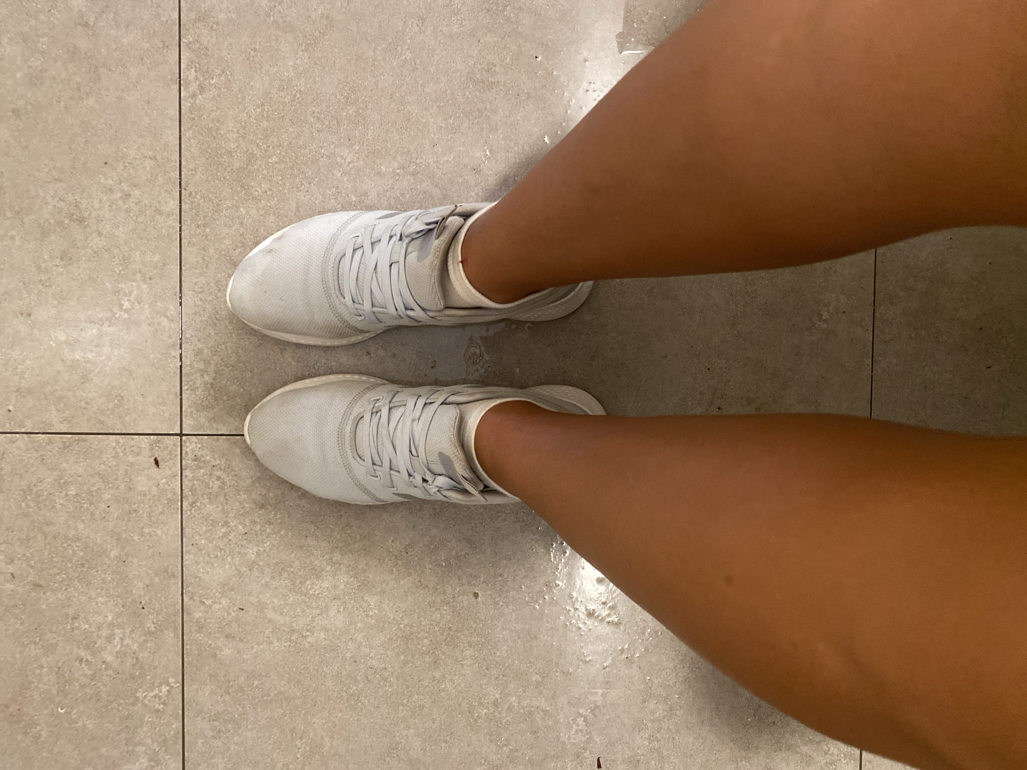 A top-down, eye-level shot shows a person's legs and feet, which are clad in white athletic shoes and white socks, standing on a light-colored tiled floor. The tiles are arranged in a grid pattern, with visible grout lines. There are small dark specks, possibly dirt or debris, scattered across the tiles. Some areas of the tile appear wet or shiny, indicating a possible spill. The person's legs are tanned and bare, extending from just below the knees down to the ankles. The shoes are tied with white laces and appear to be a mesh material. The lighting is somewhat dim, casting soft shadows. This scene does not provide any visual clues to determine its location within the city of Aksu, Turkey.