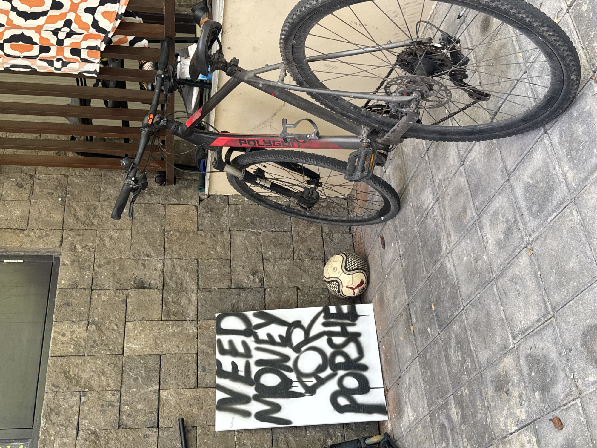 Outside a building constructed of grey, irregularly shaped stone blocks, a grey and red Polygon mountain bike leans against the building's wall. A soccer ball rests on the grey stone-tiled ground near a white canvas with black spray-painted text that reads "NEED MORE MONEY FOR PORSCHE." The location appears to be in Kuta Selatan, Indonesia. A partially visible brown wooden railing and a fabric with orange, black, and white patterns are in the background.