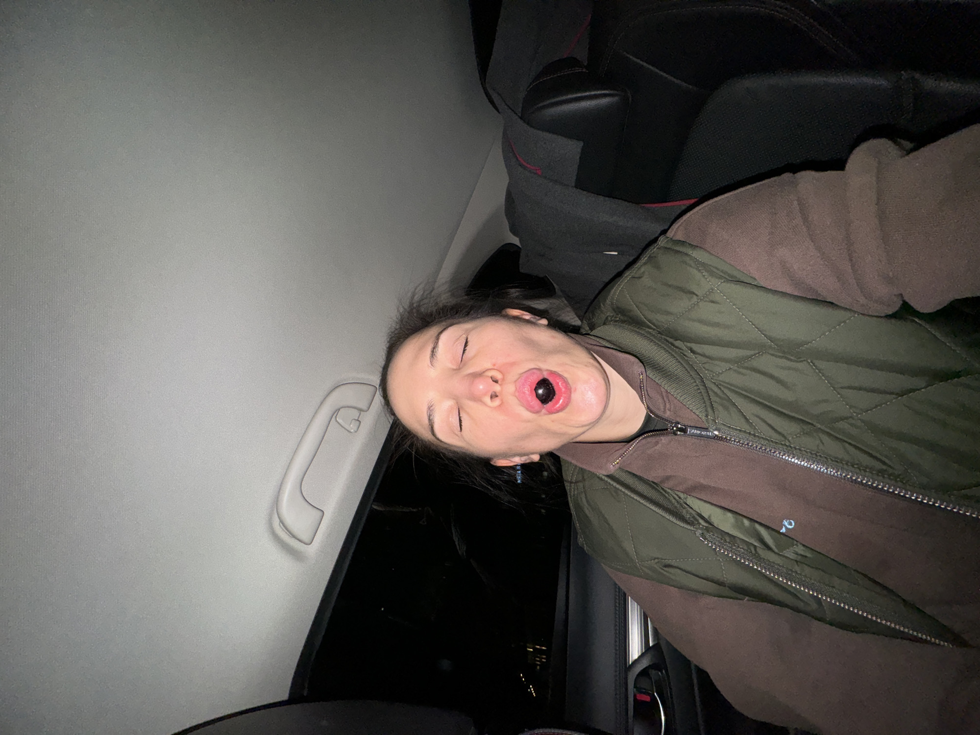 The image shows a person inside a car at night, captured from a sideways perspective. The main subject is a young person, whose face is prominent with eyes closed or squinting and mouth wide open. Inside their mouth is a large, dark, glossy, spherical object, possibly a berry or candy. They are dressed in a brown long-sleeved top, layered with a dark green quilted vest that has a metallic zipper.
The setting is the interior of a vehicle, likely a car. A light gray headliner is visible above, featuring a plastic grab handle on the left side. To the right and behind the person, a dark car seat with faint red stitching or trim is visible. The car window on the left reveals a dark exterior with indistinct, distant lights, indicating it is nighttime. The lighting within the car is dim. On the zipper pull of the green vest, there is faint, indistinct text. The person's expression appears to be exaggerated or playful. The scene is consistent with being in an urban environment like Moscow, Russia, at night.