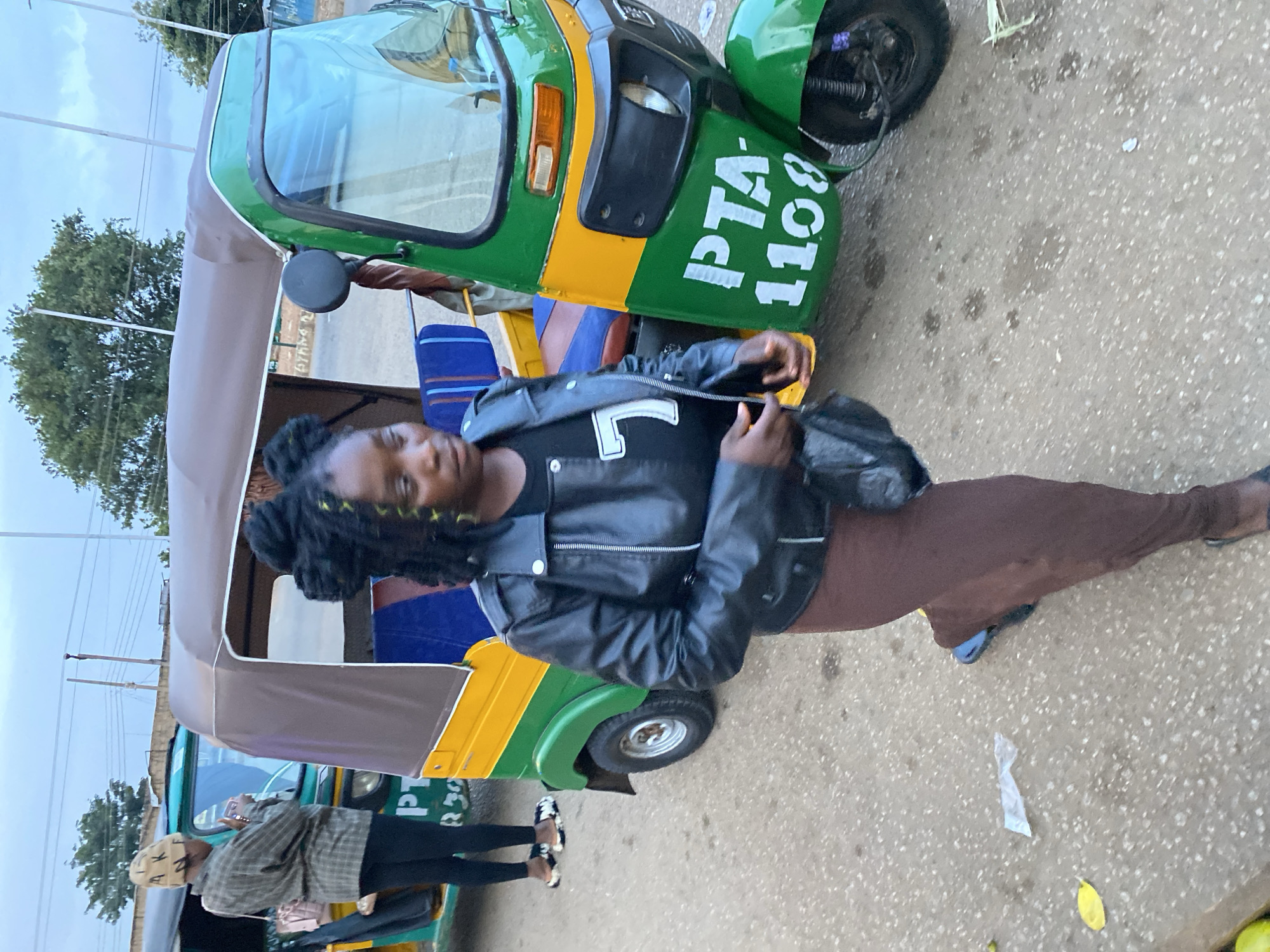 A medium shot captures a young Black woman walking past a green and yellow auto-rickshaw in Bukuru, Nigeria. The woman, facing right with her body angled towards the viewer, has black dreadlocks with a few strands of yellow. She is wearing a black leather jacket over a black t-shirt with a white "7" printed on it, and a long brown skirt. Her left hand holds a black clutch bag. The auto-rickshaw, parked on a paved street, has the white lettering "PTA 1108" on its green front panel. The background shows more auto-rickshaws and trees under an overcast sky. To the left of the main subject, another woman wearing a patterned jacket and a beige beanie is partially visible, holding a phone. The ground is a mixture of asphalt and concrete with scattered litter.
