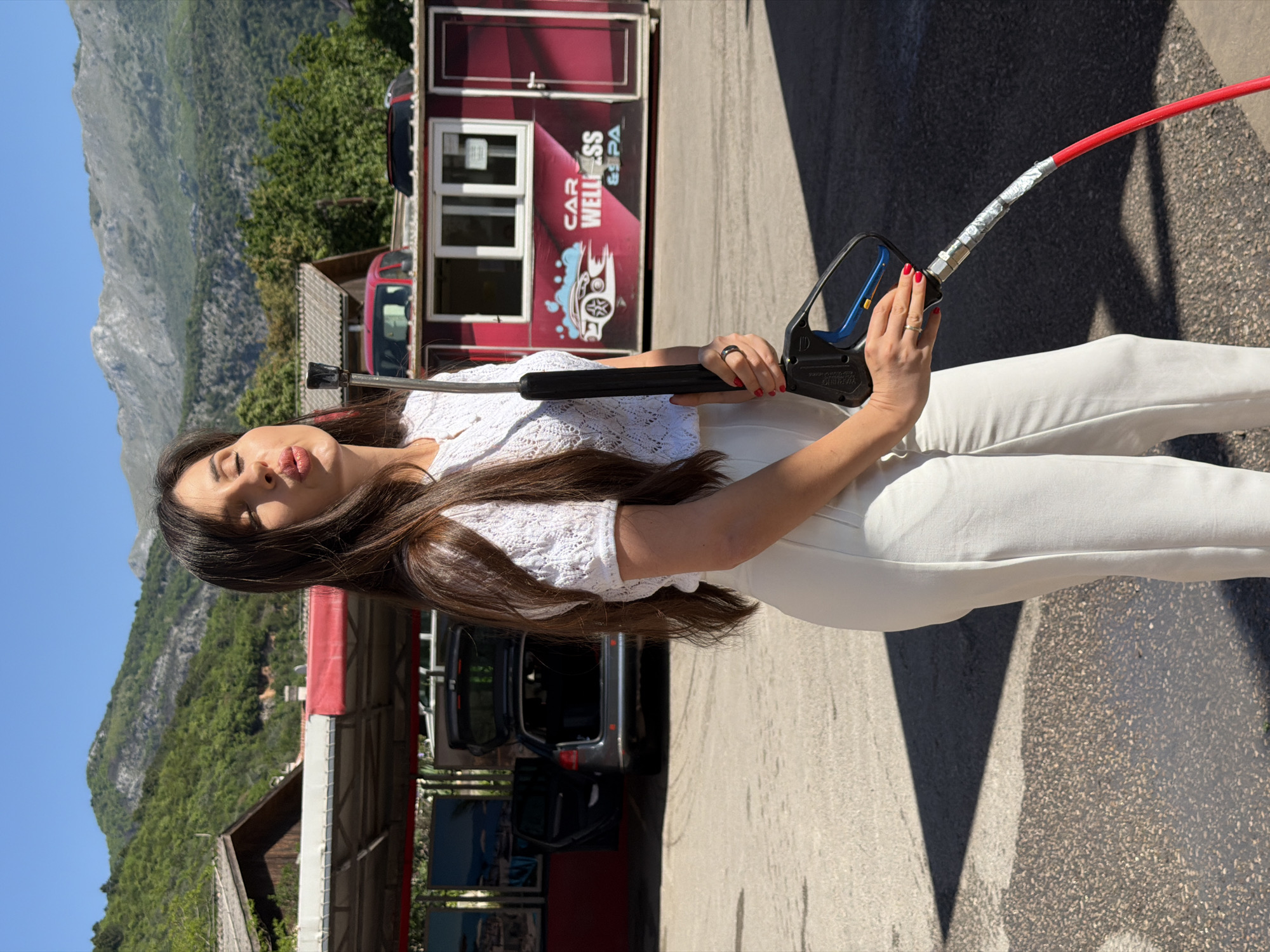 A woman is holding a pressure washer gun at an outdoor car wash. She is wearing a white knit top and white pants. Behind her, there is a car and a building with the words "CAR WELLNESS & SPA" on the side. Mountains are visible in the background. The location is likely in or near Budva, Montenegro.