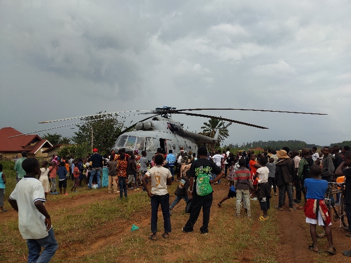 In Beni, Democratic Republic of the Congo, a large grey helicopter is landed on an unpaved, reddish-brown dirt clearing under a heavily overcast sky. The helicopter, featuring a prominent main rotor with five blades and a tail rotor, is surrounded by a crowd of approximately 50-70 individuals. The crowd consists of adults and numerous children, many of whom are observing the aircraft from various distances. Some individuals are positioned close to the helicopter's fuselage, particularly near an open side door, with a concentration of people seemingly interacting with the helicopter's interior. In the foreground, several young males stand on a dirt path, facing the helicopter. To the left background, two structures with distinct red-tiled roofs are visible, indicating a residential area. Beyond the helicopter, dense green foliage and trees form the horizon. A bicycle is present near the crowd on the right side of the frame.