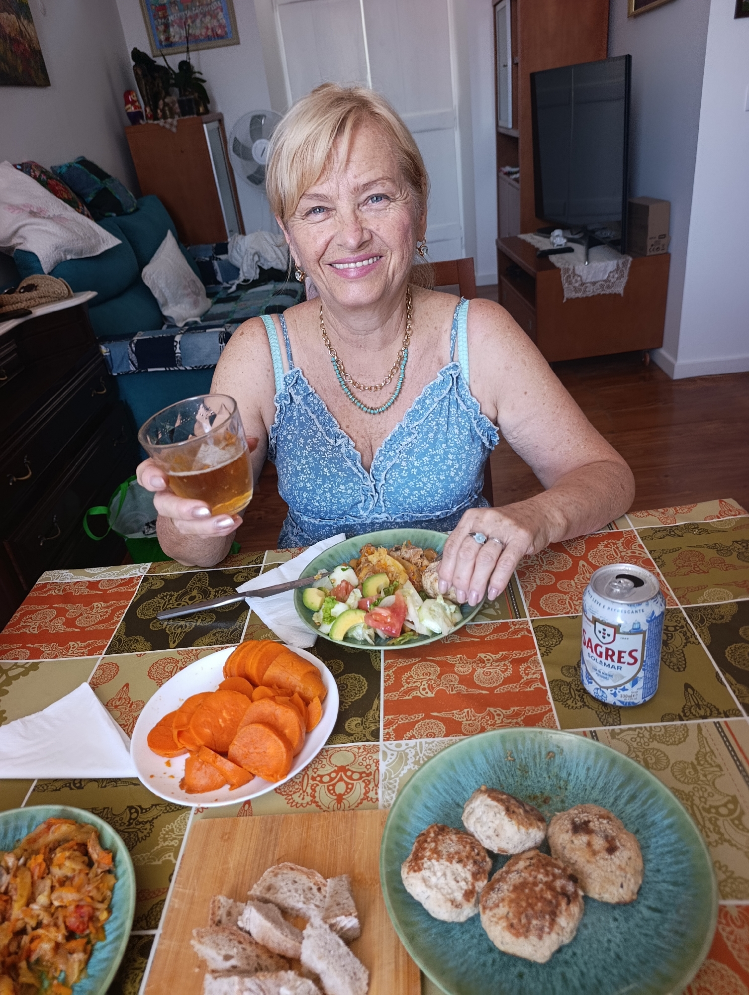 A Caucasian woman with blonde hair and blue eyes is sitting at a table and holding a glass of beer. She is wearing a blue dress and a necklace. In front of her is a plate of food and a can of Sagres beer. The table is covered with a colorful tablecloth. In the background, there is a couch and a television. The woman appears to be in a home environment.