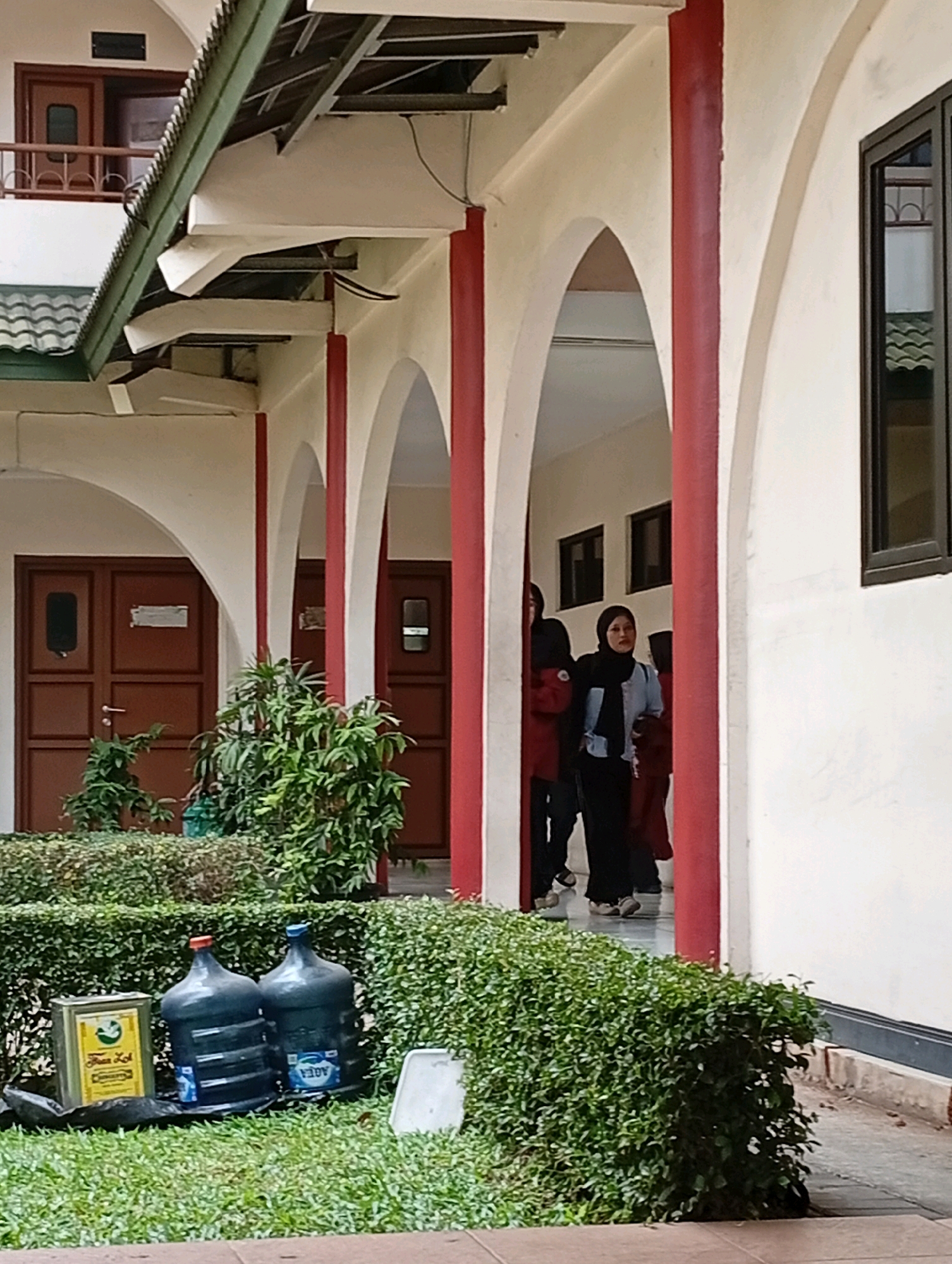 The image captures a covered walkway with red pillars and arched openings. In the foreground, a low, manicured green hedge runs horizontally. To the left, on the grass, are two large, dark blue water cooler jugs, one with a blue cap and the other with an orange cap. Beside them is a rectangular yellow container with green lettering. Behind the hedge, several green plants are visible, and further back are two brown wooden doors with rectangular glass panels and metal handles. In the background, through the arches, three individuals are walking. One person in the center wears a light blue hijab and a dark outfit, appearing to be looking towards the camera. Another person to their left wears a red top and black pants, while a third person to the right is partially obscured. The architecture suggests an institutional or educational setting, possibly a school or community building, in Kelapa Dua, Indonesia.