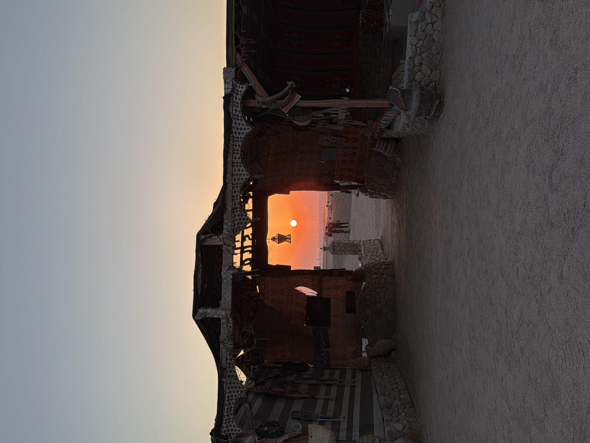 This image captures a serene sunset scene in Sharm El Sheikh, Egypt, likely near a beach or desert area. The foreground features a rustic, open-air structure resembling a tent or shelter, constructed with stone foundations and draped with fabric and woven materials. Ornate textiles and decorative elements adorn the structure, suggesting a traditional or cultural setting, possibly for tourism.
Through the central opening of this structure, the setting sun is visible as a vibrant orange orb low on the horizon, casting a warm glow over the landscape. The sky transitions from a pale blue at the top to a soft orange hue near the horizon, indicating the twilight hour. A decorative lantern hangs from the roof of the structure, silhouetted against the sunset.
In the mid-ground, beyond the shelter, several figures can be seen standing and walking, their forms partially obscured by the distance and the low light. The ground is sandy and uneven, consistent with a desert or beach environment. The overall atmosphere is peaceful and picturesque, highlighting the natural beauty of the Egyptian landscape at dusk. The structure appears to be a focal point, framing the sunset and inviting viewers to step through into the illuminated vista.
