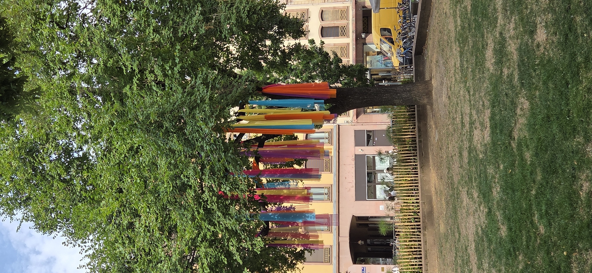 The image depicts a tree decorated with colorful fabric streamers, likely in a public space such as a park or plaza. The tree is large and leafy, providing shade. Behind the tree, several buildings with traditional European architecture are visible, characterized by yellow and pink facades and multi-paned windows. In front of the buildings, there is a low fence made of wooden posts, and beyond that, a patch of grass and a tree trunk. To the right, a yellow van is parked alongside several bicycles, suggesting a street or sidewalk nearby. A few people are seated at tables on a patio in front of one of the buildings. The sky is visible at the top of the image, with some clouds. This scene is located in Strasbourg, France, a city known for its picturesque streets and buildings.