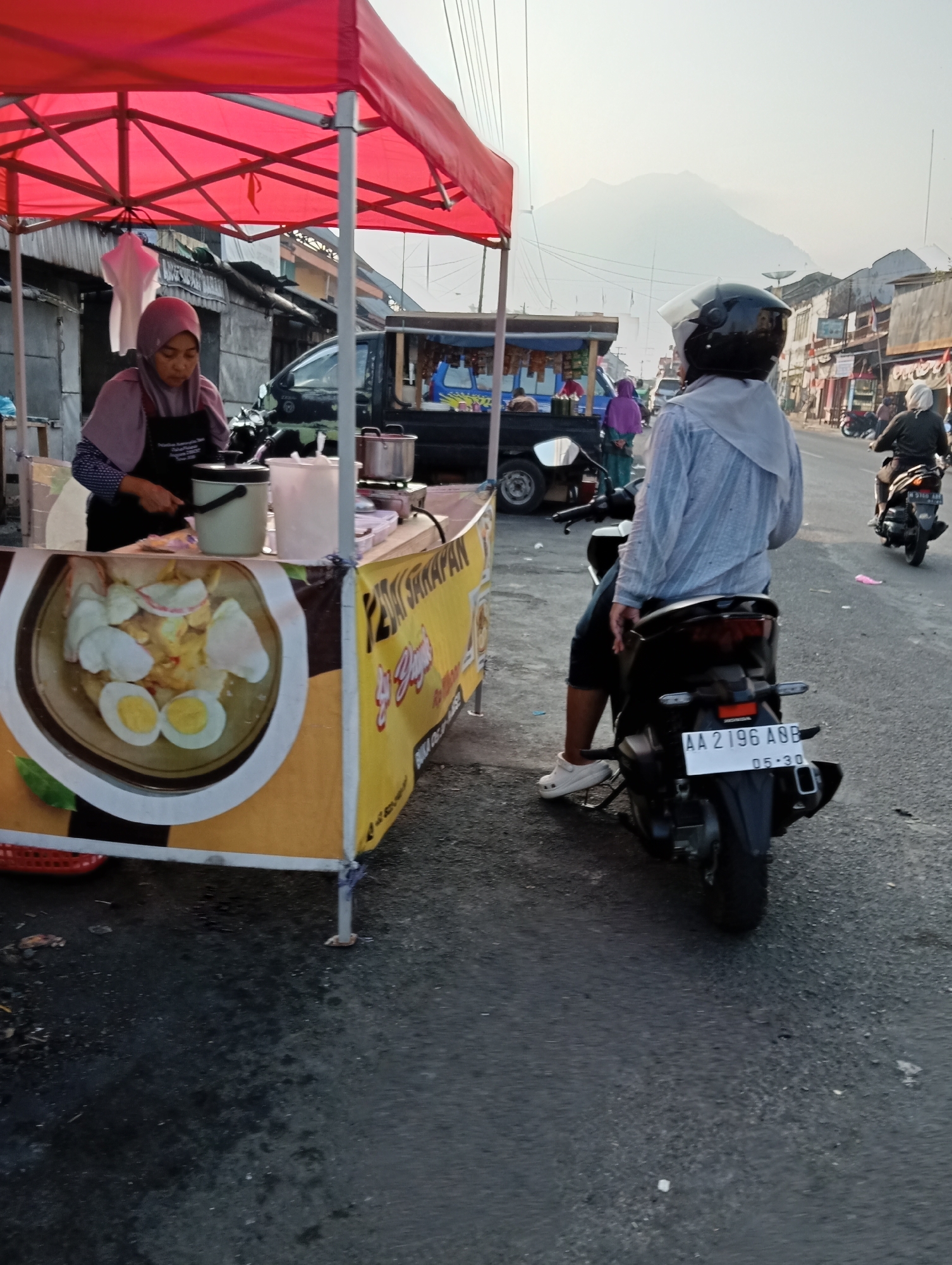 The image depicts a street scene in Grabag, Indonesia, at what appears to be a roadside food stall. In the foreground, a person wearing a light blue long-sleeved shirt and white Crocs sits on a dark-colored motorcycle, facing away from the camera. The motorcycle's license plate reads "AA 2196 ABB". To the left, a woman wearing a pink hijab and an apron stands behind a food stall with a red canopy. She is preparing food, with various containers and utensils visible. The stall's banner features a picture of a dish with eggs and crackers, and text that appears to be in Indonesian. In the background, other food stalls, buildings, a truck, and more motorcycles are visible. A large, hazy mountain can be seen in the distance, indicating a rural or semi-urban setting. The lighting suggests it might be late afternoon or early morning.