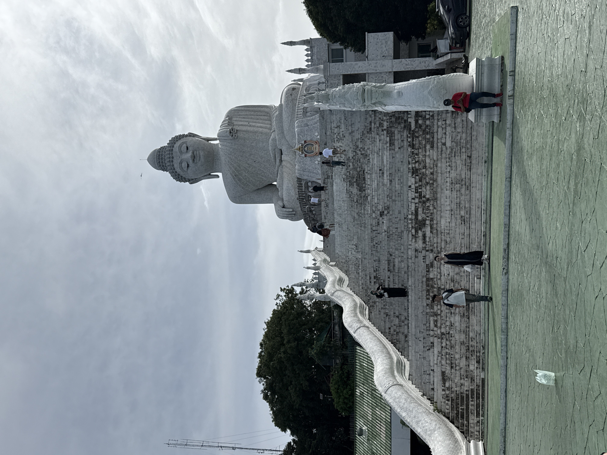 In Karon, Thailand, a large white marble statue of Buddha sits atop a hill overlooking the area. In front of the statue are a set of stairs, flanked by two white Naga sculptures on either side. Several individuals are present on the stairs and in the foreground, with some ascending the stairs towards the Buddha statue and others standing or walking on the ground level. The sky is overcast with clouds. A cell tower is visible in the background on the left side of the image.