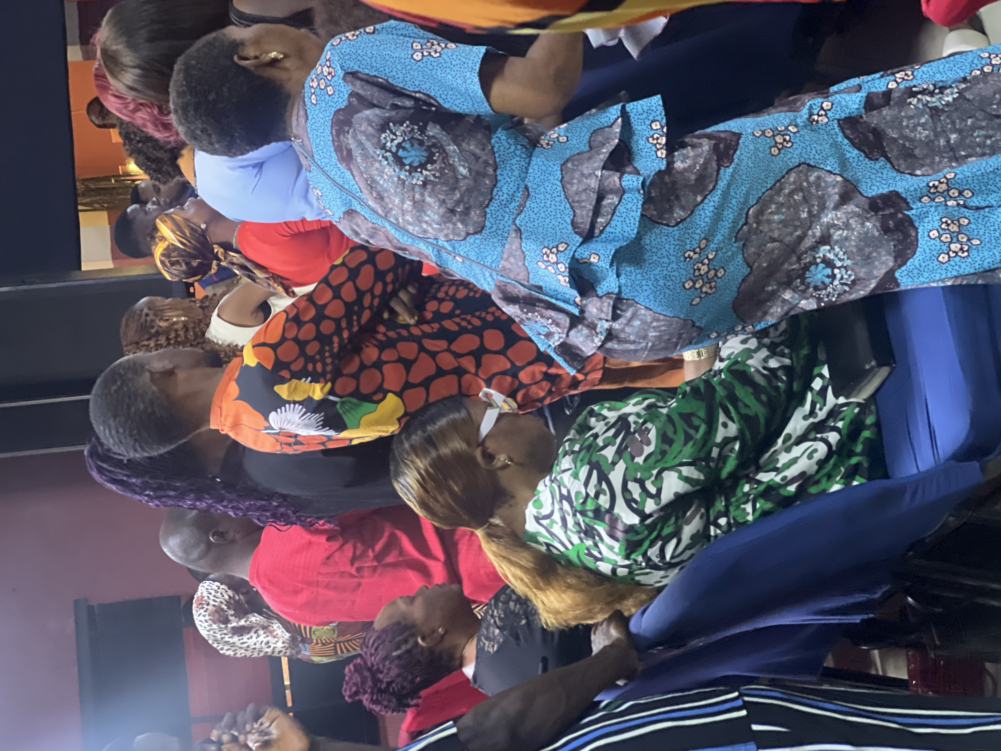 A gathering of people is visible in Nkpor, Nigeria. The attendees, mainly women, are dressed in colorful, patterned garments. The environment is dimly lit, potentially indoors, and the people seem to be observing or partaking in an event. Details like the actions, interactions, and specific objects in view are somewhat limited due to the image angle and resolution.