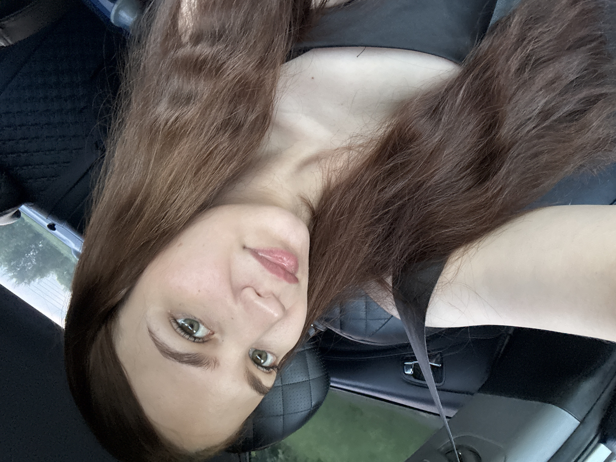 The image shows a young woman with long, brown hair, sitting in the driver's seat of a car. She is looking towards the camera with a slight smile. The interior of the car is visible, including a black leather seat and a seatbelt. The woman is wearing a black tank top. Outside the car, greenery and trees can be seen, suggesting a rural or suburban environment. The overall lighting suggests it is daytime. The location within the city of Кварса, Russia, cannot be determined from the image alone.