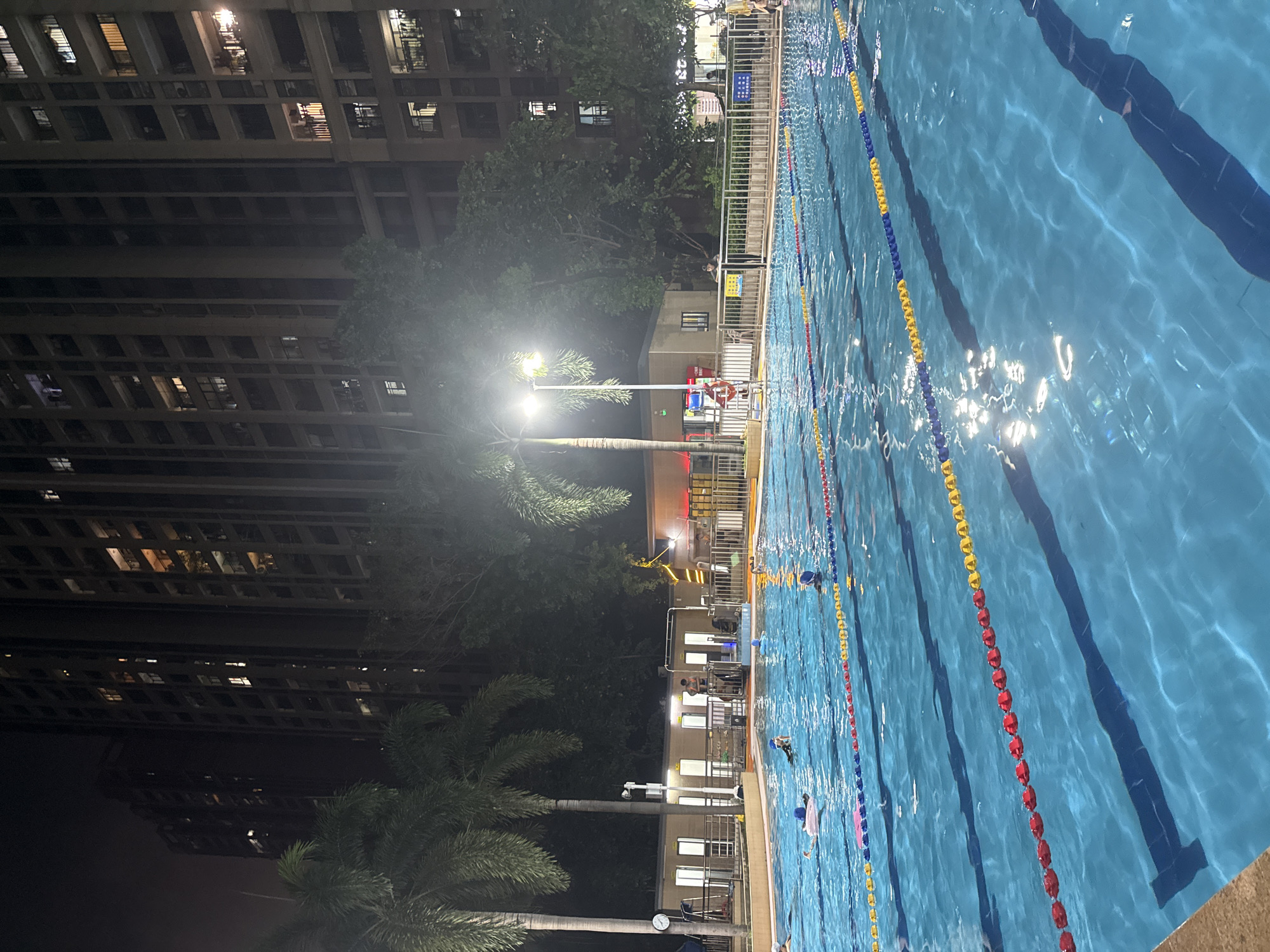 The image depicts an outdoor swimming pool at night, illuminated by overhead lighting and the lights from surrounding buildings. Tall residential buildings are visible in the background, suggesting an urban setting. Palm trees are present on the left side of the image. Several people are in the pool, some swimming and others in the shallow end. Lane dividers are visible in the pool, indicating it is set up for lap swimming. A building with multiple rooms or cubicles is situated behind the pool, and a fence encloses the pool area. The water is clear blue and reflects the light. The overall atmosphere is one of a recreational facility in operation after dark. The specific location is confirmed to be in Haizhu District, China.