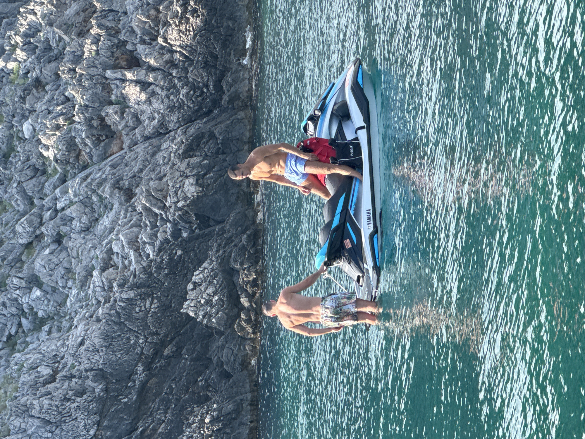 Two men are on a Yamaha jet ski in the clear, blue-green waters off the coast of Dubraljevina, Montenegro. The sun is shining brightly, illuminating the choppy water. One man, wearing blue patterned swim trunks, stands waist-deep in the water, holding onto the jet ski. He has a muscular build and appears to be enjoying the day. The other man, wearing red swim trunks and blue patterned shorts, sits on the back of the jet ski. He has a towel draped over his shoulders. The jet ski is white with blue accents and has "YAMAHA" written on the side. In the background, a steep, rocky cliff face rises from the water, covered in sparse vegetation. The time of day appears to be late morning or early afternoon, given the strong sunlight and the shadows cast by the rocks. The scene conveys a sense of leisure and adventure.