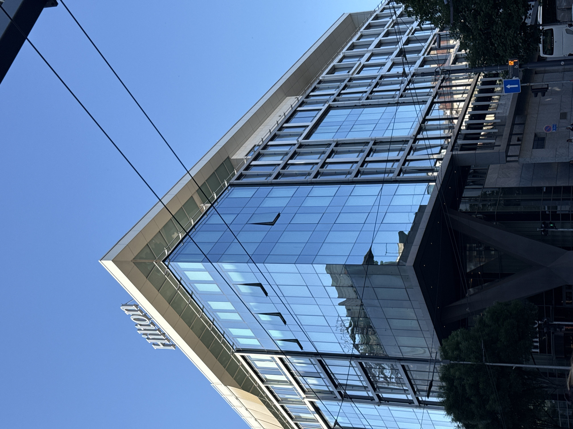View of the Hilton Hotel in Belgrade, Serbia, specifically within the Vracar Urban Municipality. The building is modern, with a facade of large glass panes and a grid-like structural design. The hotel's name, "Hilton," is visible on the lower portion. Some windows are open. Utility lines run across the top left of the frame. The sky is clear and blue. Reflected in the glass is the surrounding urban landscape. A street sign and traffic lights are visible.