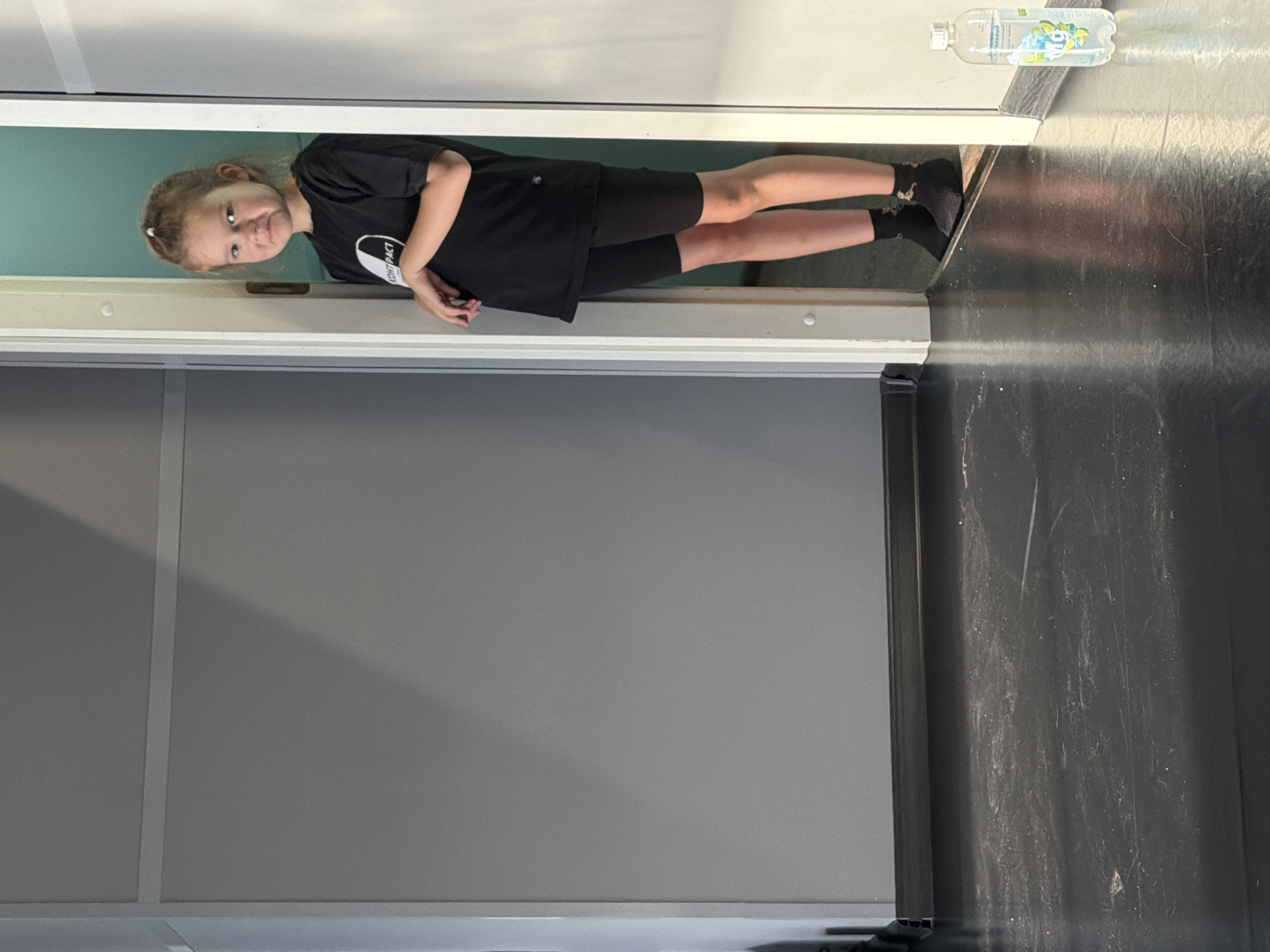 A young girl with blonde hair is standing in the doorway of a room. She is wearing a black t-shirt and black shorts. The room has gray walls and a black floor. A water bottle is on the floor near her feet. The image does not provide enough information to determine the location in the city of Saint Petersburg, Russia.