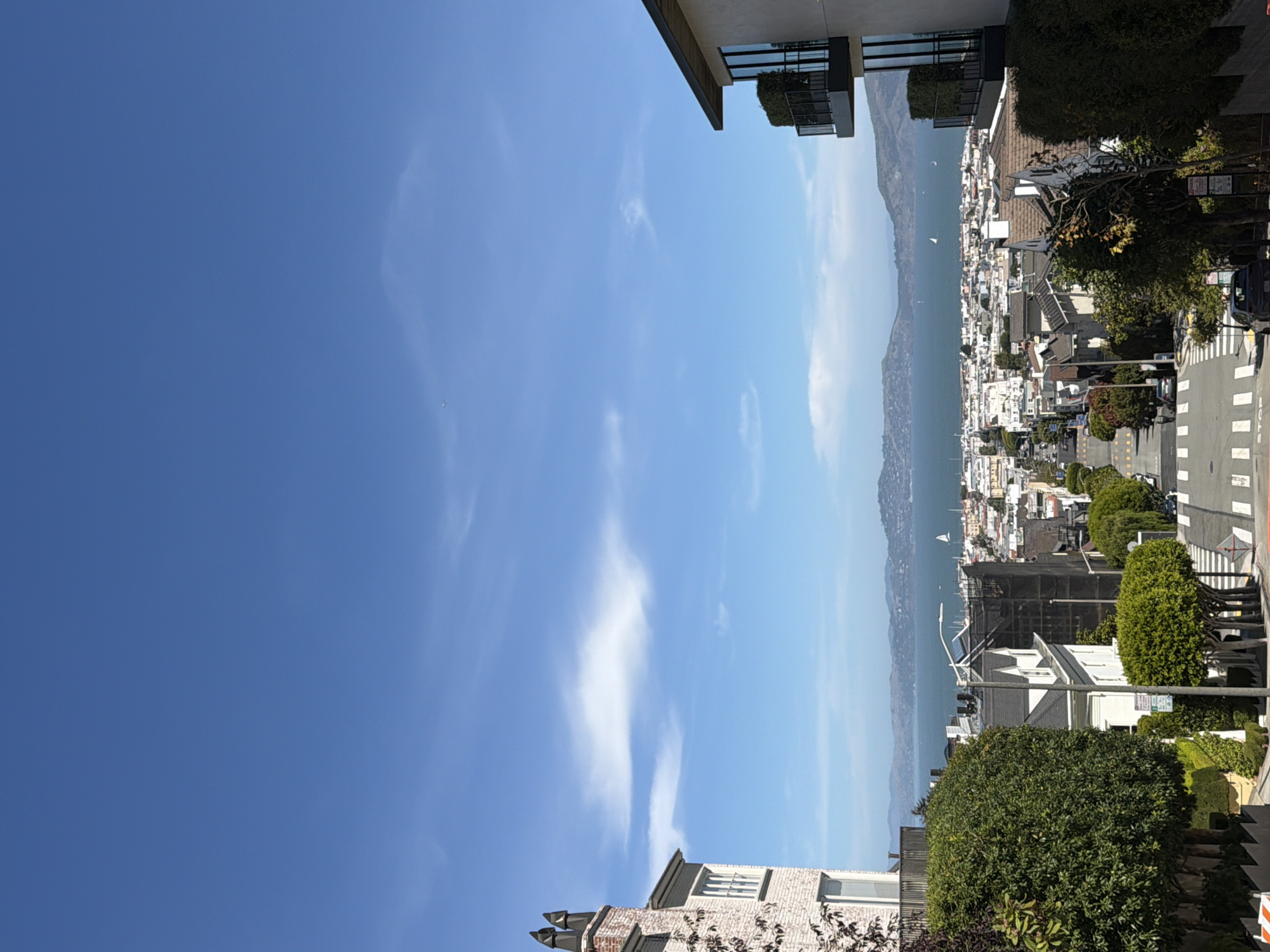 This is a photograph taken from a high vantage point in San Francisco, California, looking down a steep residential street towards the San Francisco Bay. The sky is clear and blue with scattered white clouds.
In the foreground, on the left, is a portion of a light-colored brick building with multiple windows and two distinctive chimney tops. On the right, a modern building with balconies and large windows is visible.
The street itself is paved with asphalt and marked with white crosswalks. It descends steeply into the distance. Several cars are parked along the street and a few are in motion. A pedestrian is visible walking down the street. Residential houses with varying architectural styles line both sides of the street, many featuring pitched roofs and trees in front. Green, manicured trees and bushes are abundant throughout the scene.
In the middle ground and background, a dense urban landscape of buildings stretches towards the water. Beyond the city, the expansive San Francisco Bay is visible, with sailboats dotting its surface and hills forming the horizon. The overall impression is a classic San Francisco vista, characterized by its hilly terrain, diverse architecture, and proximity to the bay.