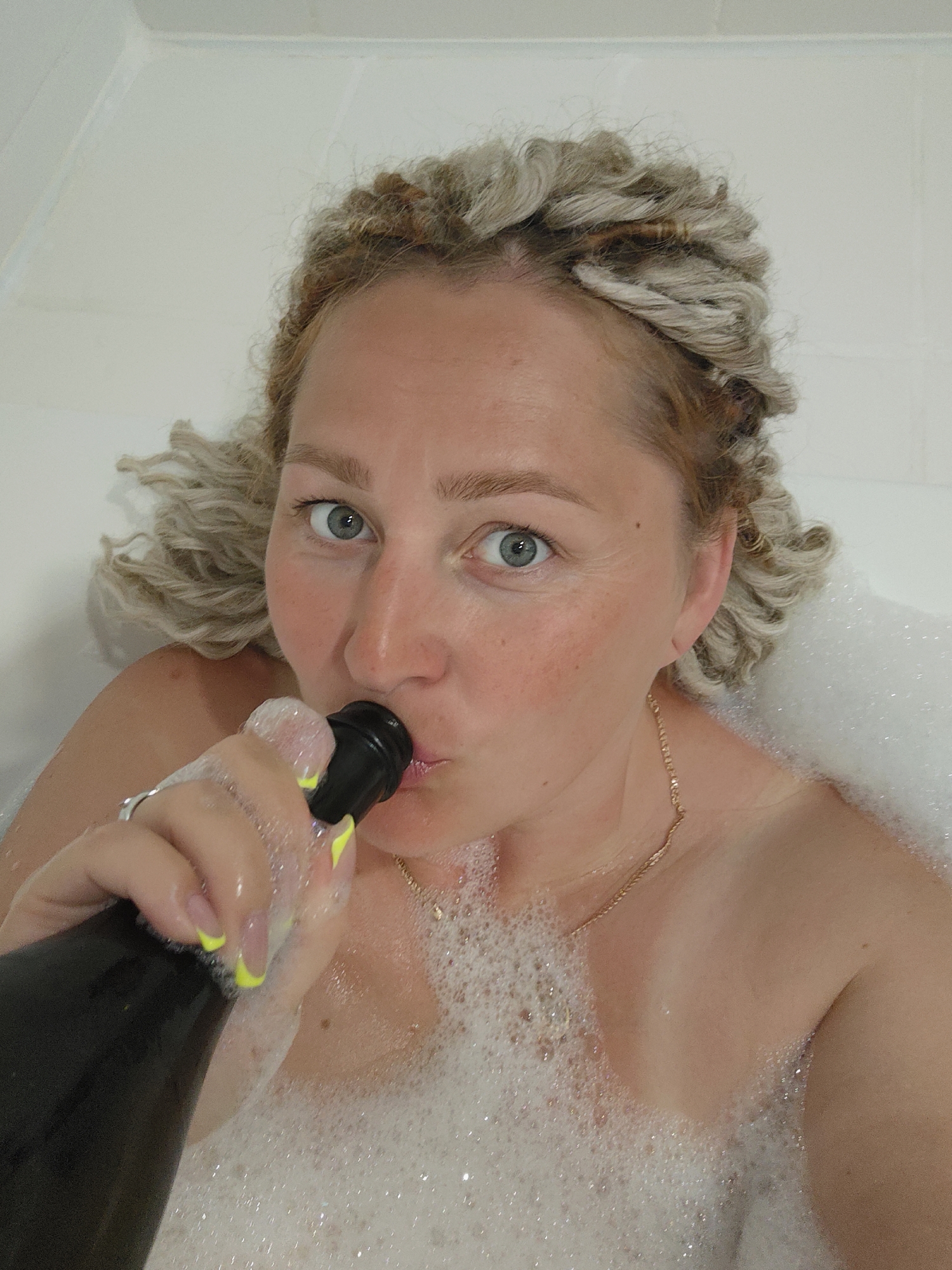 A woman is taking a selfie in a bathtub filled with bubbles. She is holding a dark bottle with a black cap to her mouth, appearing to drink from it. Her hair is styled in blonde dreadlocks, and she is wearing a gold necklace. Her fingernails are painted with a French manicure and neon yellow tips. The environment is a white bathtub. The image does not contain any recognizable landmarks or features that would allow for identification of a specific location within the city of Sochi, Russia.