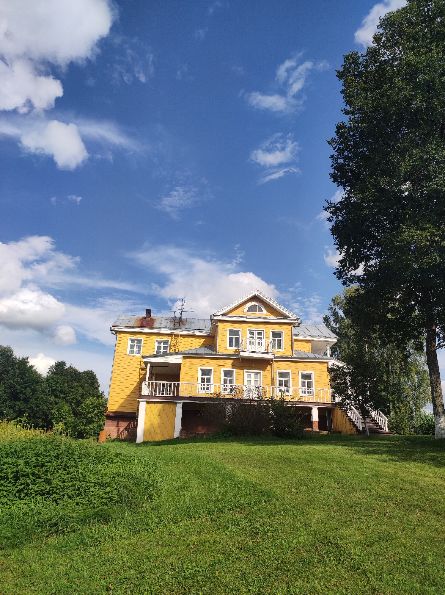 A bright yellow, two-story house with a gray roof stands on a grassy hill under a blue sky with white clouds. The house has white-trimmed windows and balconies with white railings. A large tree with dark green leaves is to the right of the house. The ground in front of the house is covered with green grass and some overgrown bushes. The location is Zavod Mikhailovskii, Russia.