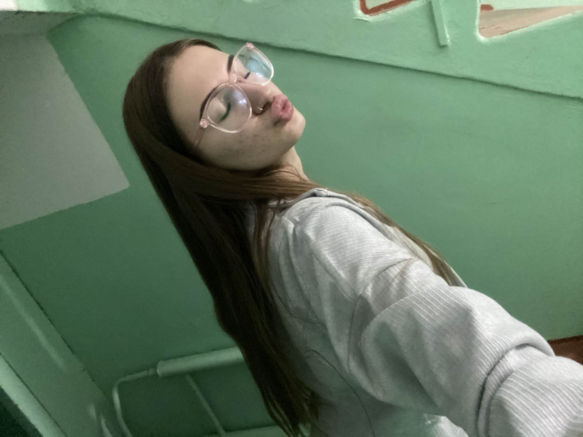 A low-angle, interior photograph captures a young female with long, dark brown hair, looking upwards and to her right, in the city of Unknown, Unknown. She wears clear-framed, light pink cat-eye glasses, with her eyes closed or squinting, and her lips puckered into a "kiss face." A small stud piercing is visible on her left nostril. Her complexion shows minor blemishes on her cheeks. She is dressed in a light grey, ribbed or textured, long-sleeved hooded sweatshirt. The background is predominantly a solid, matte mint green wall. In the upper right, a raised horizontal architectural feature with an irregular reddish-brown outline is visible. The upper left shows a diagonal junction of two green surfaces. In the lower left, a white linear object, possibly a pipe or railing, is present against a darker green section. The lighting appears to be artificial and diffused.