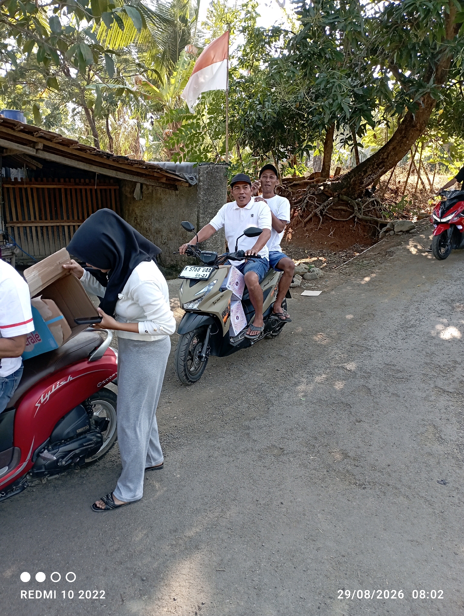 In Dukuhseti, Indonesia, on August 29, 2026, at 8:02 AM, a scene unfolds near a weathered structure with a wooden fence. An Indonesian flag waves from a pole. A woman wearing a black hijab and a white long-sleeved shirt is seen interacting with a cardboard box, which rests on the seat of a red scooter. She is also holding a phone. Beside her, a man in a white polo shirt and blue shorts, accompanied by another man, sits on a beige scooter. The beige scooter has a license plate that reads "K 6768 UAA 06-23" and is adorned with decorative flags. In the background, a red motorcycle is parked. The environment is characterized by trees, foliage, and a dirt and gravel road.