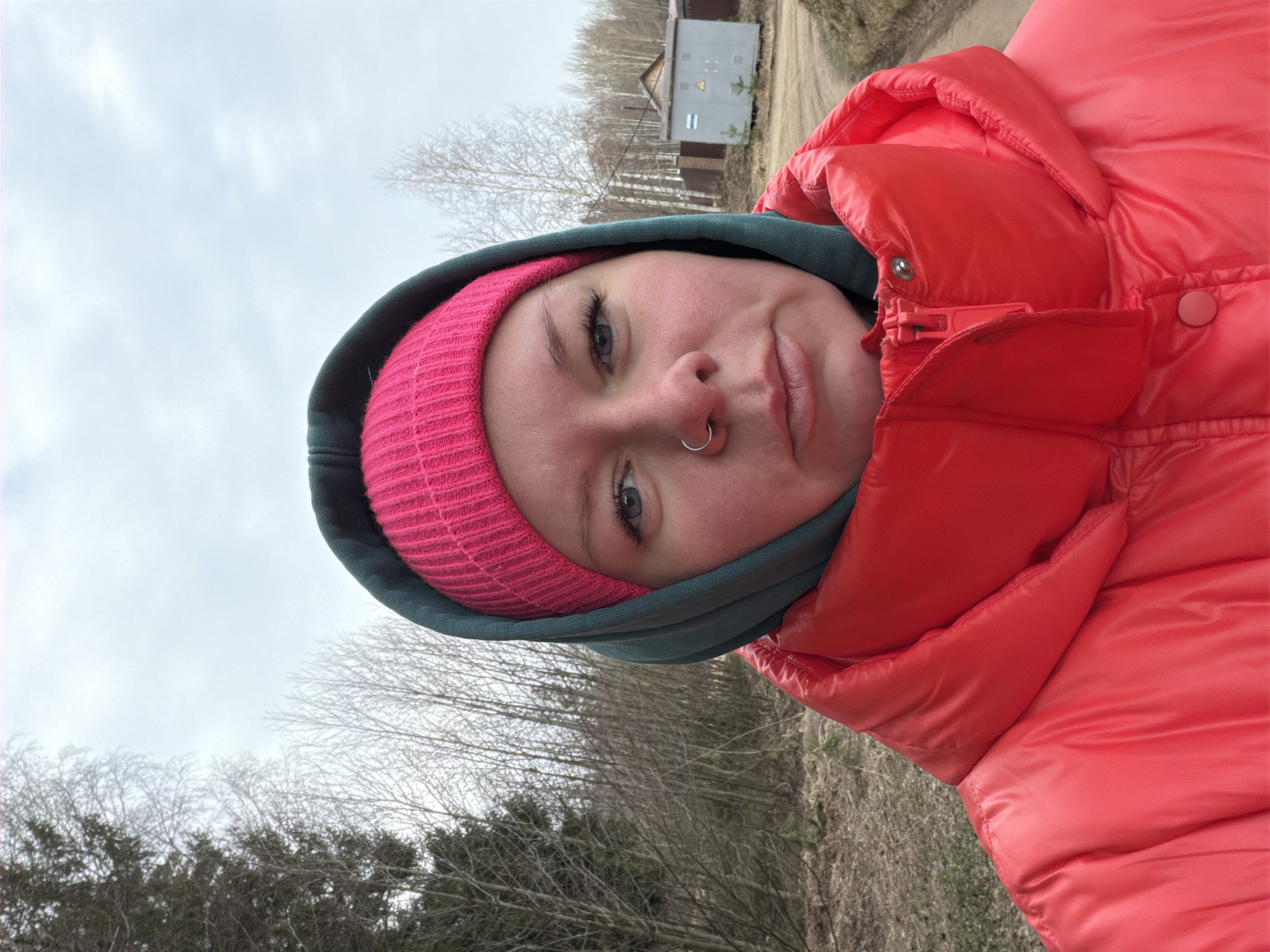 A person is wearing a pink beanie under a green hood and a red puffer jacket outdoors. The background shows trees without leaves, a dirt path, and a small utility building. The location is the city of Chaikovsky, Russia.