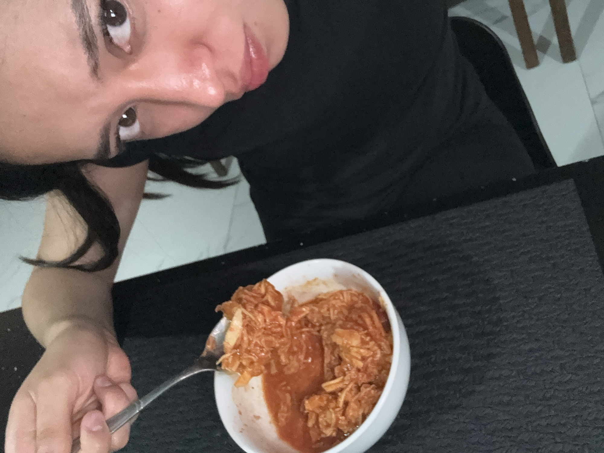 A person wearing black clothing is holding a fork with a spoonful of orange-colored food from a white bowl. The bowl is filled with more of the same food and some liquid. The person is sitting at a dark gray textured table. The background shows a white tiled floor and the leg of a chair. The location is unknown; there is nothing in the image to indicate that it is in Puebla, Mexico.