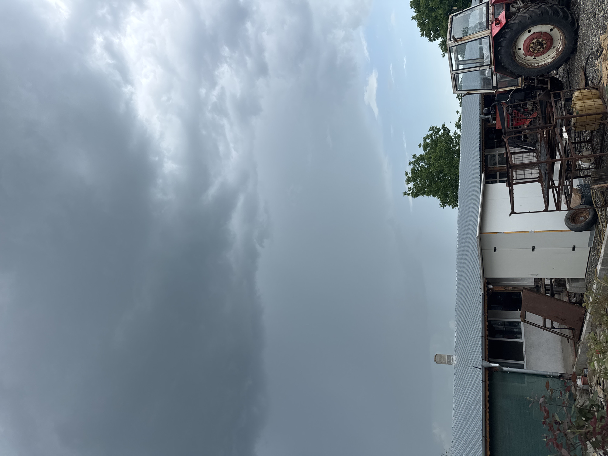 The image shows a rural scene in Sarnevo, Bulgaria. A large portion of the sky is filled with dark, ominous clouds, indicating a potential storm. To the right of the image, a section of a red tractor is visible, with a large, worn tire. Beside it are rusted metal carts.
Adjacent to the tractor is a section of a low-slung, single-story building. The building has a gray roof, white walls, and visible windows. A green awning covers a portion of the structure.
Vegetation, including trees, is sparsely scattered in the background. The scene suggests a farm or rural property on a day with threatening weather.