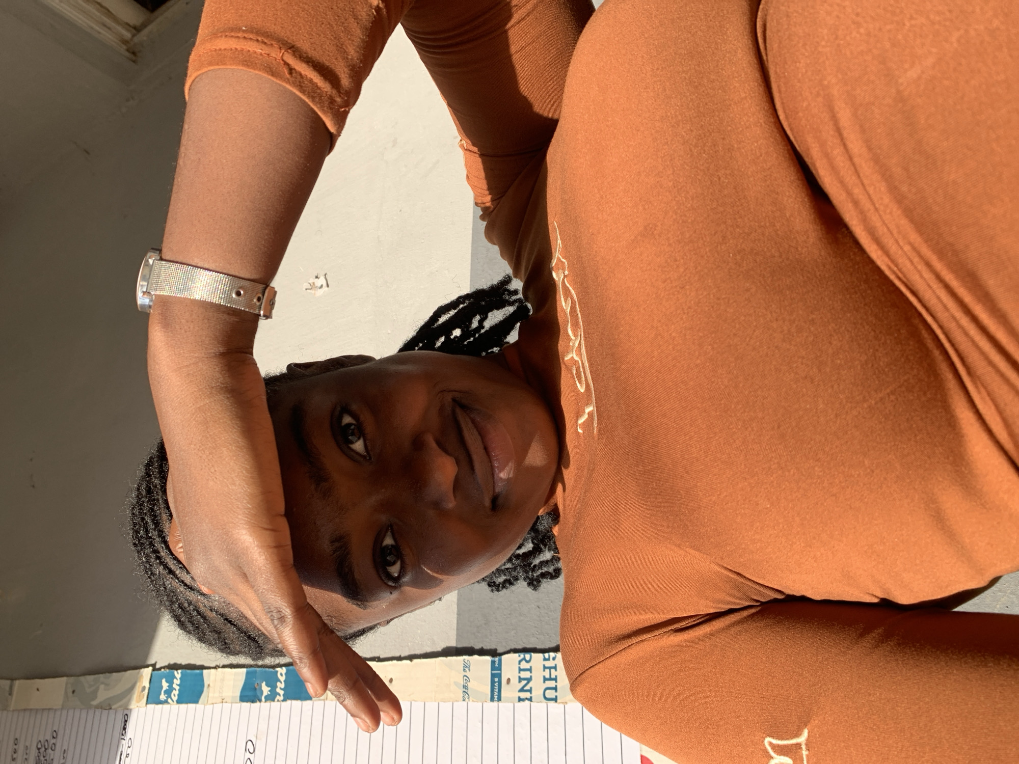 A dark-skinned individual with braided hair, wearing a form-fitting, long-sleeved, burnt orange top with white embroidered script on the chest and a partial letter on the left arm, looks directly into the camera. A silver-colored mesh strap watch is visible on her left wrist. Her right hand is raised, palm outwards, shielding her eyes from a strong, overhead light source, which casts a distinct shadow across her forehead and upper face. The background consists of a light-colored wall to which multiple sheets of paper, containing handwritten numbers and text (e.g., "1500", "500"), are vertically affixed. Below these papers, a faded blue and white patterned material with inverted text (e.g., "The Coca-Cola", "SUTTAK", "RINI", "SHU") is partially visible. The scene is brightly illuminated by natural light, casting pronounced shadows. This event is occurring in New Stadium, Nigeria.