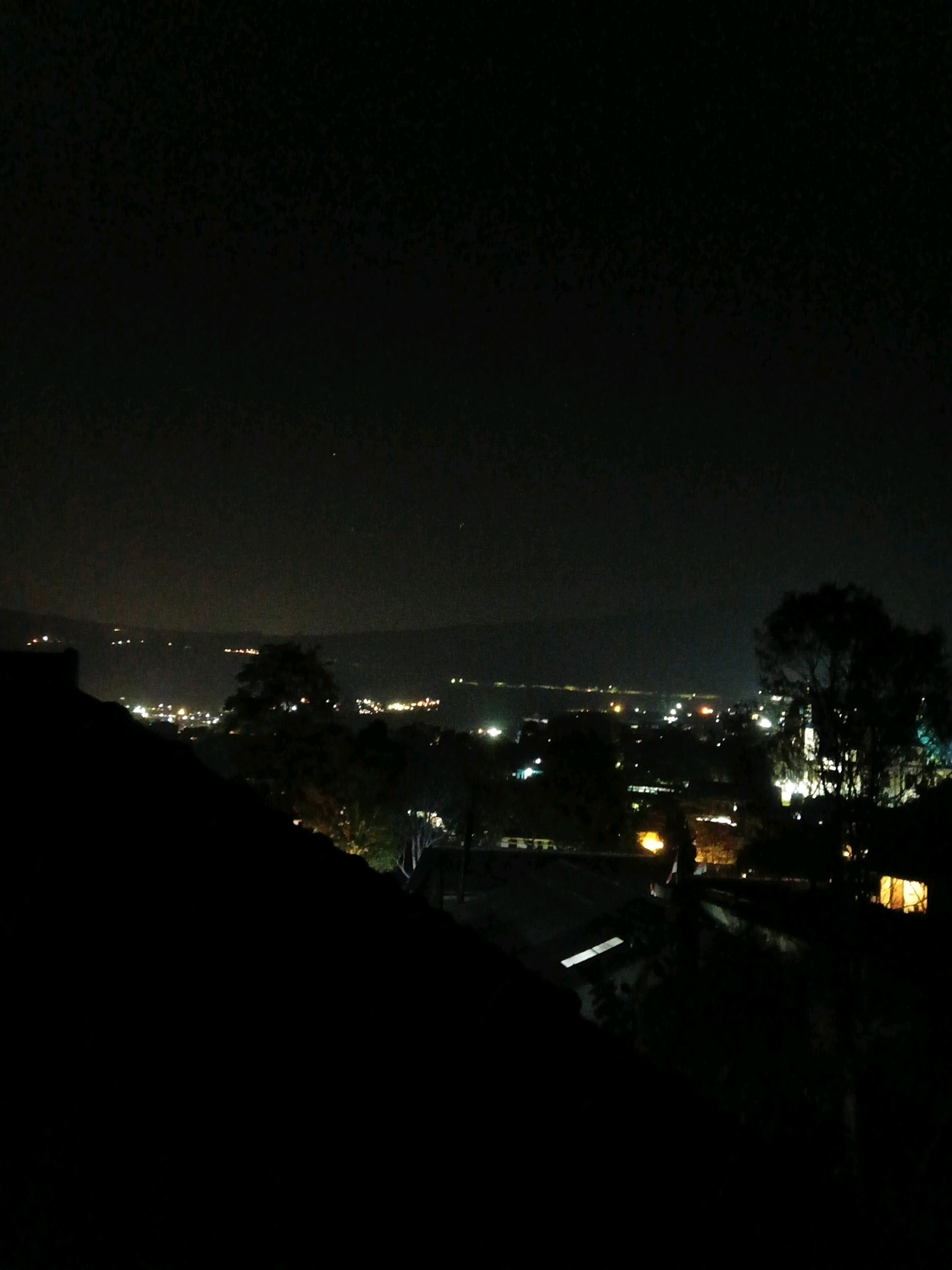 The image depicts a nighttime view of a city, likely Ciater, Indonesia, seen from an elevated position. The foreground is dominated by a dark, silhouetted roofline, possibly part of a building or structure, which slopes downwards from the left side of the frame. Beyond this, the midground reveals a sprawling urban landscape illuminated by numerous lights. These lights form patterns indicative of streets, buildings, and possibly homes, stretching into the distance. Several trees with dark foliage are scattered throughout the midground and background, their shapes discernible against the illuminated city. The background consists of a dark sky, with faint lights suggesting distant settlements or possibly hills. One notable feature in the midground is a single building with a warmly lit window, standing out against the darker surroundings. The overall impression is that of a peaceful, yet active, urban environment at night.