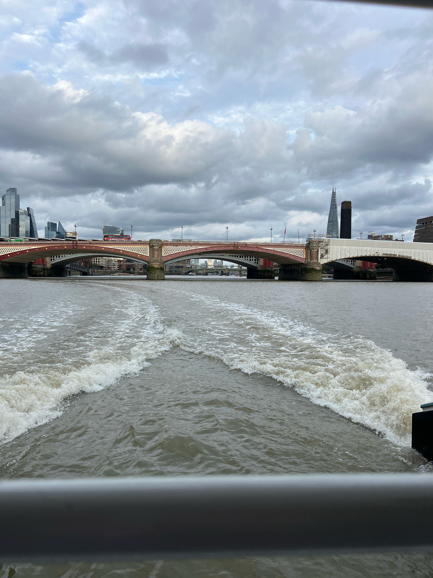 The image shows a view from a boat on the River Thames in London, United Kingdom. In the foreground, the wake of the boat creates foamy white waves. In the midground, a large bridge with red and white accents spans the river. Several other bridges are visible in the distance, including one with a distinctive white arch. The London skyline is visible in the background, featuring modern skyscrapers like the Shard and older buildings. A red double-decker bus is seen crossing the bridge. The sky is overcast with dramatic grey clouds.