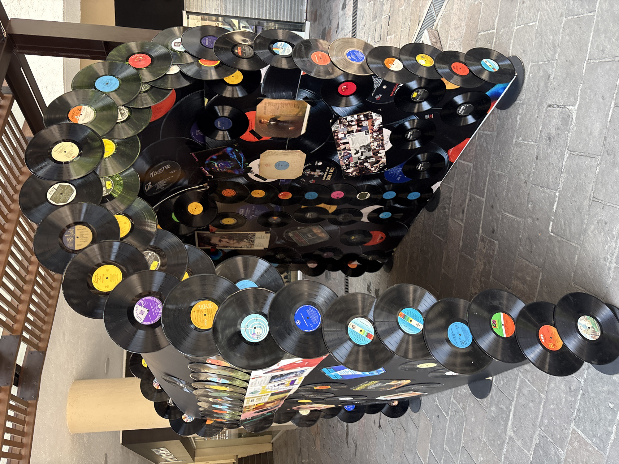 The image shows a large, arched sculpture constructed from numerous vinyl records. The sculpture is located in an outdoor setting, possibly a public walkway or plaza, with cobblestone paving visible beneath it. In the background, there is a building with a light-colored pillar and part of a wooden slatted ceiling or awning. The sculpture itself is a complex arrangement of black vinyl records of various sizes, some with colorful labels, adhered to a dark, curved structure. The records are overlapping and arranged in layers, creating a textured and visually dynamic surface. Some records are positioned with their labels facing outwards, while others are seen from their dark surfaces. The arrangement forms an archway, suggesting it is a passage or decorative feature. There are no people visible in the image. The lighting suggests it is daytime, with natural light illuminating the scene. The overall impression is one of a creative art installation using vintage music media. This sculpture is located in the city of Cuernavaca, Mexico.