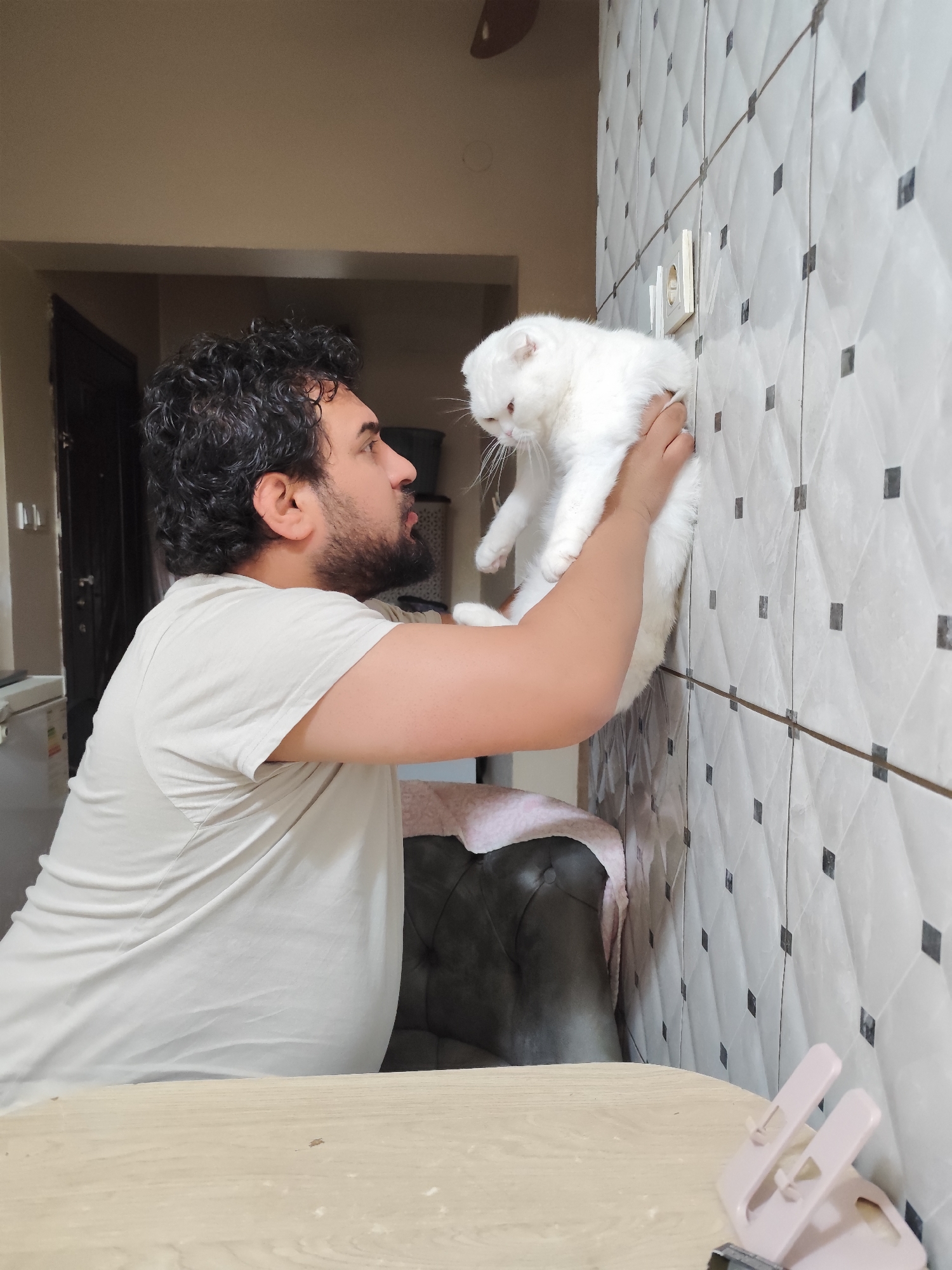 A person with dark curly hair, a beard, and a light-colored t-shirt is holding a white cat up towards a tiled wall. The cat appears to be a Scottish Fold breed, based on the appearance of folded ears. The wall is covered in white tiles with dark accents. A light beige table is in the foreground. A portion of a dark upholstered chair or sofa is visible behind the person. The location is presumed to be indoors. There is no information to verify the location is specifically Sarıyer, Turkey.
