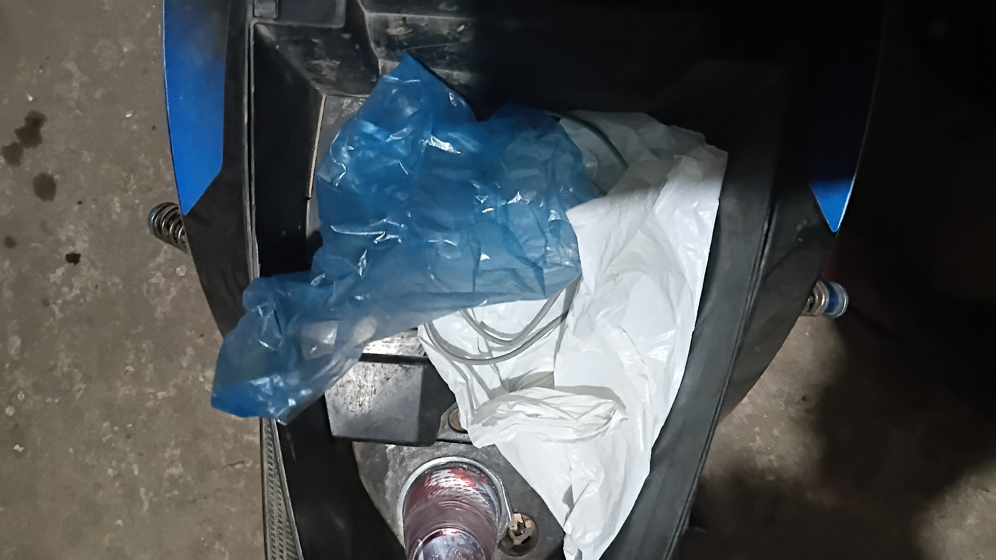 An overhead view captures the interior of a motorcycle's storage compartment. Within the compartment, a crumpled blue plastic bag rests on top of a white plastic bag. A silver-colored cable is partially visible between the two bags. To the left, a clear plastic cup with a red rim is positioned near the edge of the compartment. The compartment itself appears to be dark, with hints of blue plastic visible on the exterior. The surrounding environment suggests an outdoor setting, possibly a street or parking area, with a concrete surface visible. There are no people or any indication of specific actions or interactions. The scene is static and offers no clues about the location in Prabumulih, Indonesia.