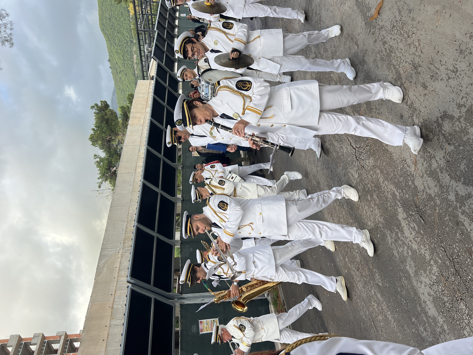 A line of musicians in white uniforms plays instruments in Budva, Montenegro. The musicians are standing in a row on a concrete surface next to a building under construction. The band includes clarinet, trumpet, saxophone, cymbals, and percussion. Mountains are visible in the background. The sky is overcast.
