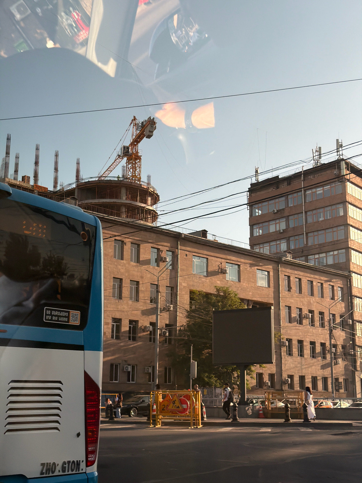 A daytime, eye-level, medium shot captures a street scene in Yerevan, Armenia, dominated by a blue and white bus in the foreground. The bus's rear is visible, displaying a license plate or identifier reading "ZHO:GTON:" and a sticker with Armenian text and a QR code. Behind the bus, a construction site is evident with a large yellow crane and unfinished building structures reaching towards a clear sky with reflections of the interior of a vehicle on the sky. Several multistory buildings, one significantly taller than the others, line the street. The buildings are constructed of light brown brick and feature numerous windows, some with air conditioning units. Overhead, a network of power lines crisscrosses the scene. On the street, pedestrians are visible, including a woman in a white dress walking to the right, and two people standing near a yellow construction barrier with signage. The street surface is asphalt, with shadows cast by the buildings and the overhead structures. A large, dark digital billboard is mounted on a pole on the sidewalk. The overall impression is an active urban environment with ongoing development.