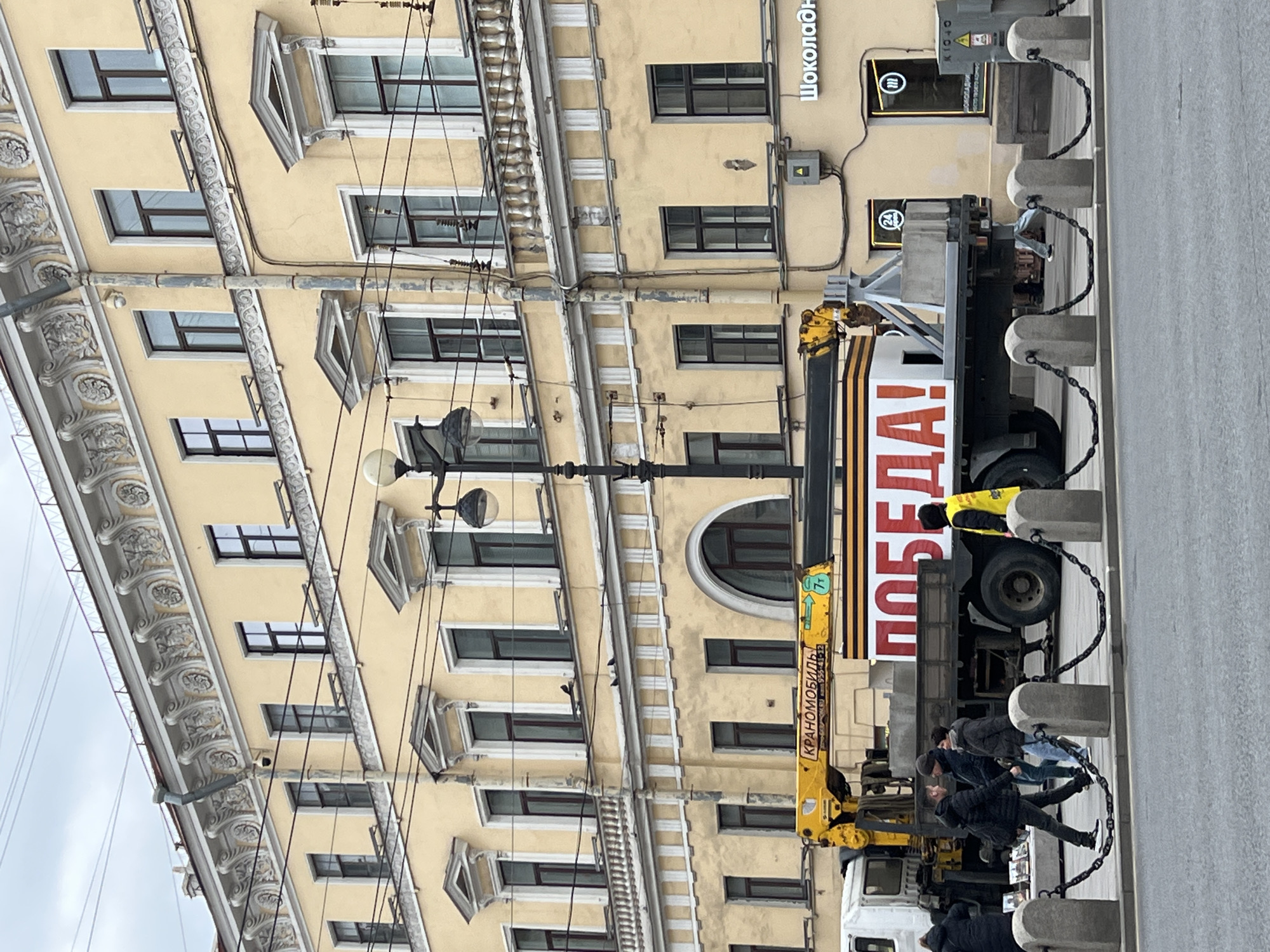 This image captures a street scene in Saint Petersburg, Russia, under an overcast sky. The prominent subject is a light yellow, multi-story building featuring classical architectural elements such as ornate white cornices, decorative window frames, and pilasters. Numerous windows are visible across its facade, along with utility wires and drainpipes.
In the foreground, a large truck, identifiable as a crane truck by its yellow extending arm labeled "КРАНОМОБИЛЬ" (Cranemobile), is parked along the street. A significant feature of the truck is a large banner mounted on its side, proudly displaying the Russian word "ПОБЕДА!" (VICTORY!) in bold red letters on a white background, accompanied by the distinctive orange and black stripes of a St. George ribbon.
Several people are visible on the sidewalk next to the truck. Some individuals are walking, while one person in a bright yellow jacket stands near the rear of the vehicle. The sidewalk is demarcated by grey concrete bollards linked by chains. Signs on the building include partial text "Шоколадн" (likely "Шоколадница," a coffee house) and a "24" symbol, indicating 24-hour service. The overall setting suggests a public space, possibly during a commemorative event, given the "Victory" banner, strongly associated with Victory Day celebrations in Russia.