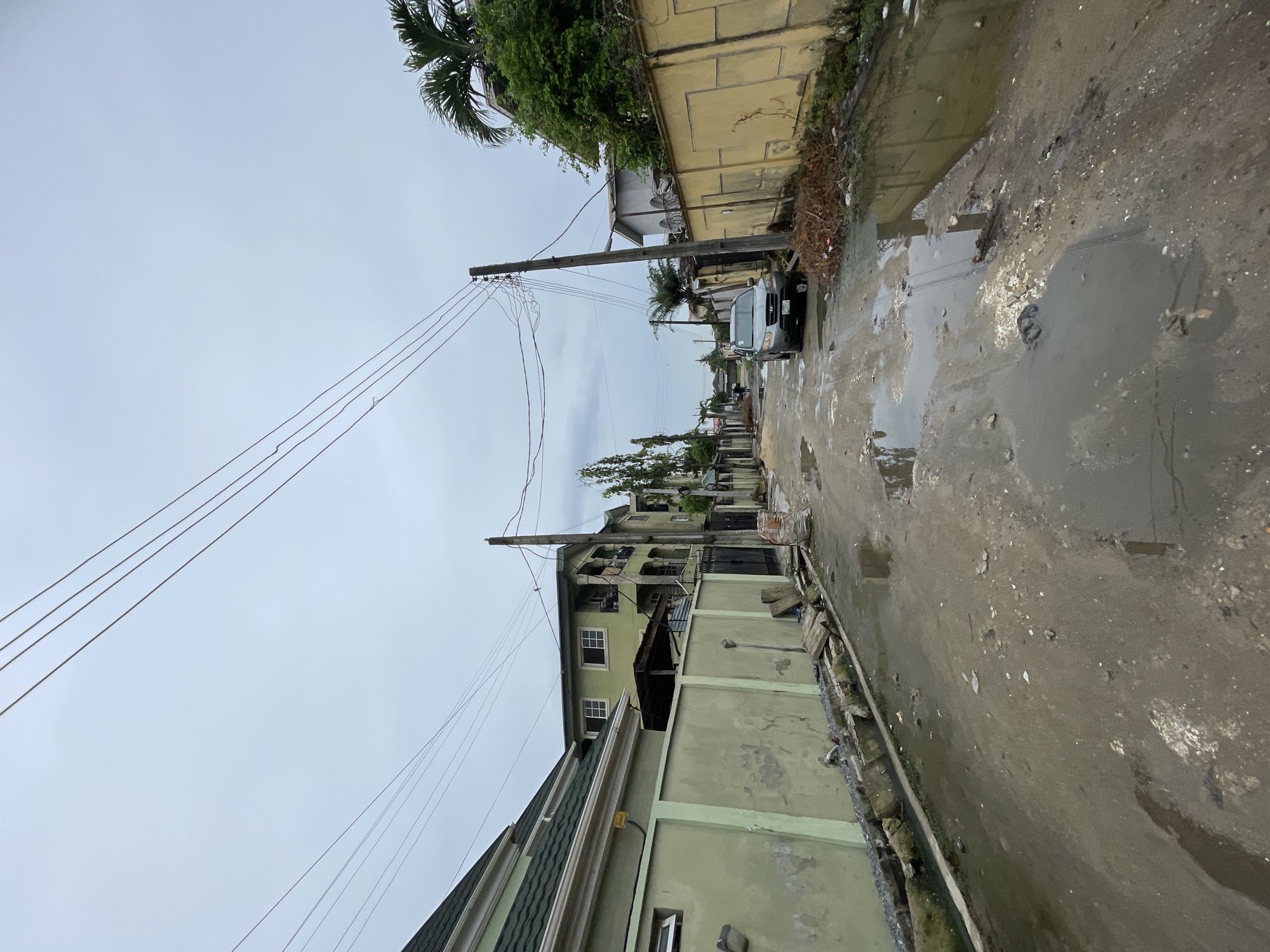 The image depicts a street in Ado, Nigeria, after rainfall, with standing water in potholes. Buildings are visible on both sides of the street, made of concrete blocks, some painted. Utility poles line the street with power lines overhead. A silver minivan is parked further down the street. The sky is overcast. The environment appears to be a residential area with signs of neglect.