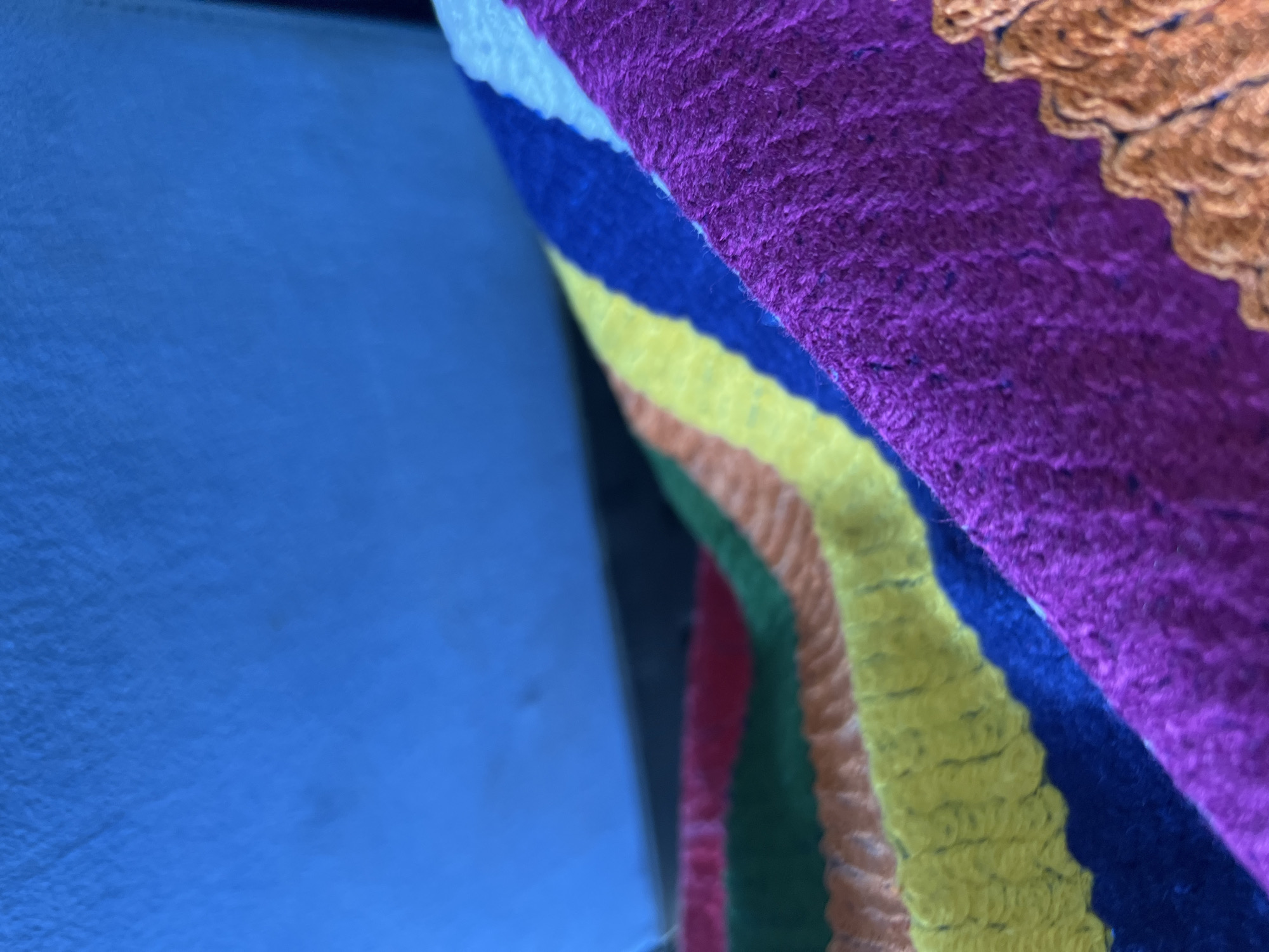 A close-up photograph taken in the city of Slivovo, Bulgaria, features a vibrant, multi-colored textile occupying the lower and mid-ground against an out-of-focus, solid blue background. The textile, possibly a blanket or shawl, exhibits distinct horizontal stripes of woven or crocheted material, showcasing colors including purple, dark blue, yellow, orange, green, and a glimpse of red. The intricate texture of the yarn is visible, particularly in the purple and yellow segments, suggesting a handcrafted item. The perspective is a shallow depth of field, rendering the blue background as an indistinct surface, likely a wall or upholstered furniture. No individuals or specific actions are depicted.