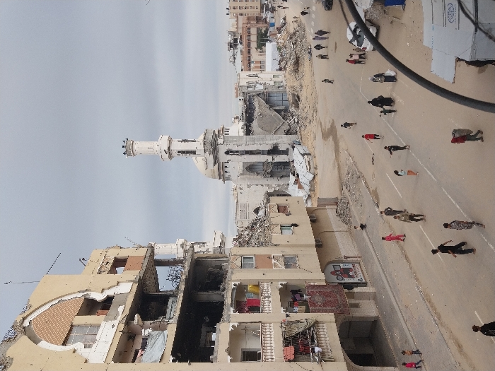 This image depicts a wide street scene in Khan Yunis, Palestinian Territory, characterized by significant urban destruction. On the left, a multi-story, beige apartment building shows extensive damage, with large sections of its facade and internal structures exposed, and windows missing. Clothing hangs on balconies, one of which also displays a red rug. In the background and center, a prominent white mosque with a tall minaret and a dome is heavily damaged, with portions of its structure collapsed and rubble visible around its base. Numerous piles of rubble are scattered throughout the scene, particularly to the right of the damaged apartment building and near the mosque.
The paved street, marked with a faded white dividing line, is occupied by approximately two dozen civilians of varying ages, including men, women, and children. They are walking in different directions; some individuals walk alone, others in small groups. One child in a pink outfit appears to be running or playing. Another adult figure is seen carrying a large, light-colored bundle on their back. A white car is parked on the right side of the street further down, and what appears to be a dark-colored vehicle is partially visible in the far right foreground. Makeshift structures, possibly tents or stalls, are also visible on the far right. The sky is overcast.