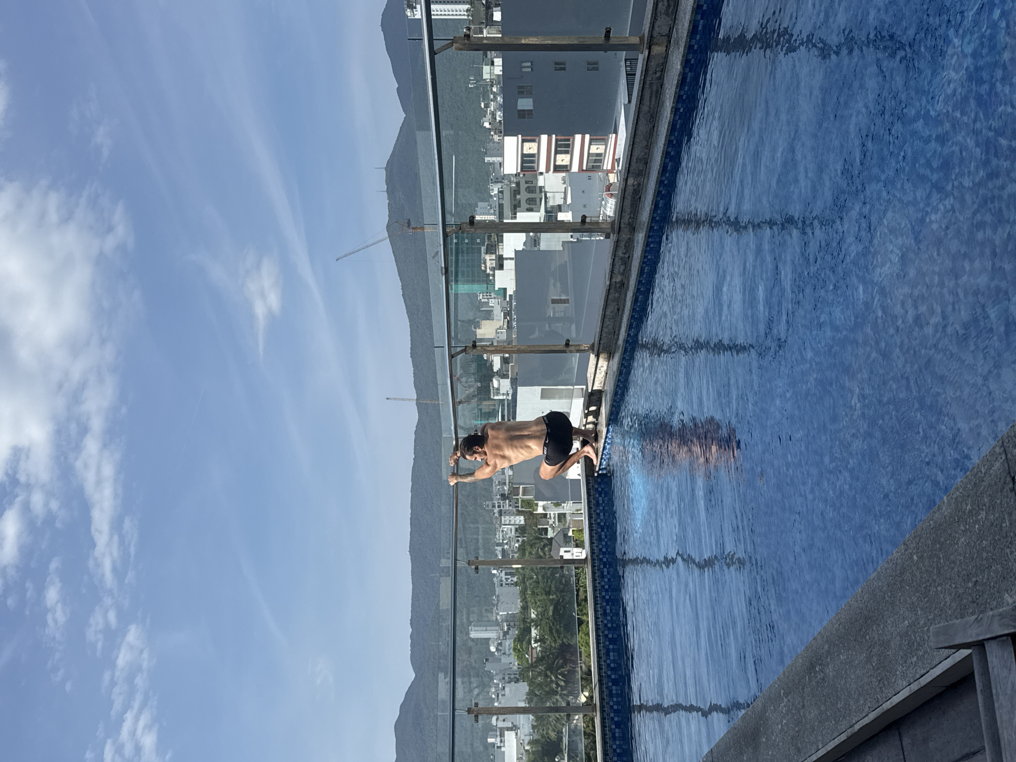 A man in black swimming trunks is hanging onto the edge of an outdoor swimming pool, preparing to enter. The pool has a light blue tiled bottom and a clear glass safety barrier. The pool appears to be on a rooftop, offering a view of the city of Đà Nẵng, Vietnam, with buildings and mountains visible in the background. The sky is clear with some scattered clouds. A construction crane is visible in the distance.