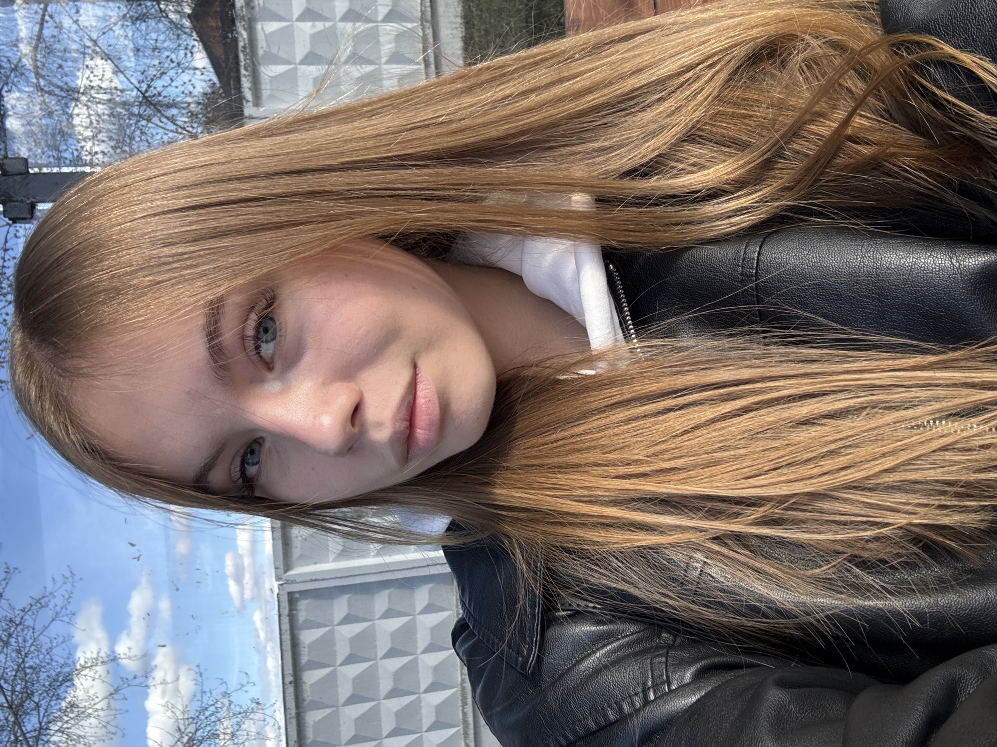 A young woman with long, light brown hair is posing for a selfie. She is wearing a black leather jacket over a white hoodie. The background includes a fence with a geometric pattern, trees without leaves, and a cloudy sky. The location may be Chaikovsky, Russia based on provided information, but this cannot be verified from the image alone.