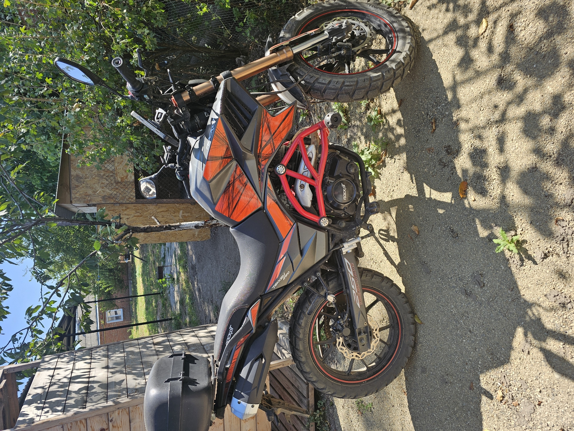 In Novoshcherbinovskaya, Russia, a black and orange dirt bike is parked on a gravelly area next to a wooden structure. The motorcycle has a black top box, a red crash cage, and knobby tires. The background shows green foliage, a fence, and a portion of a building with a white-framed window. The sun casts shadows on the ground.