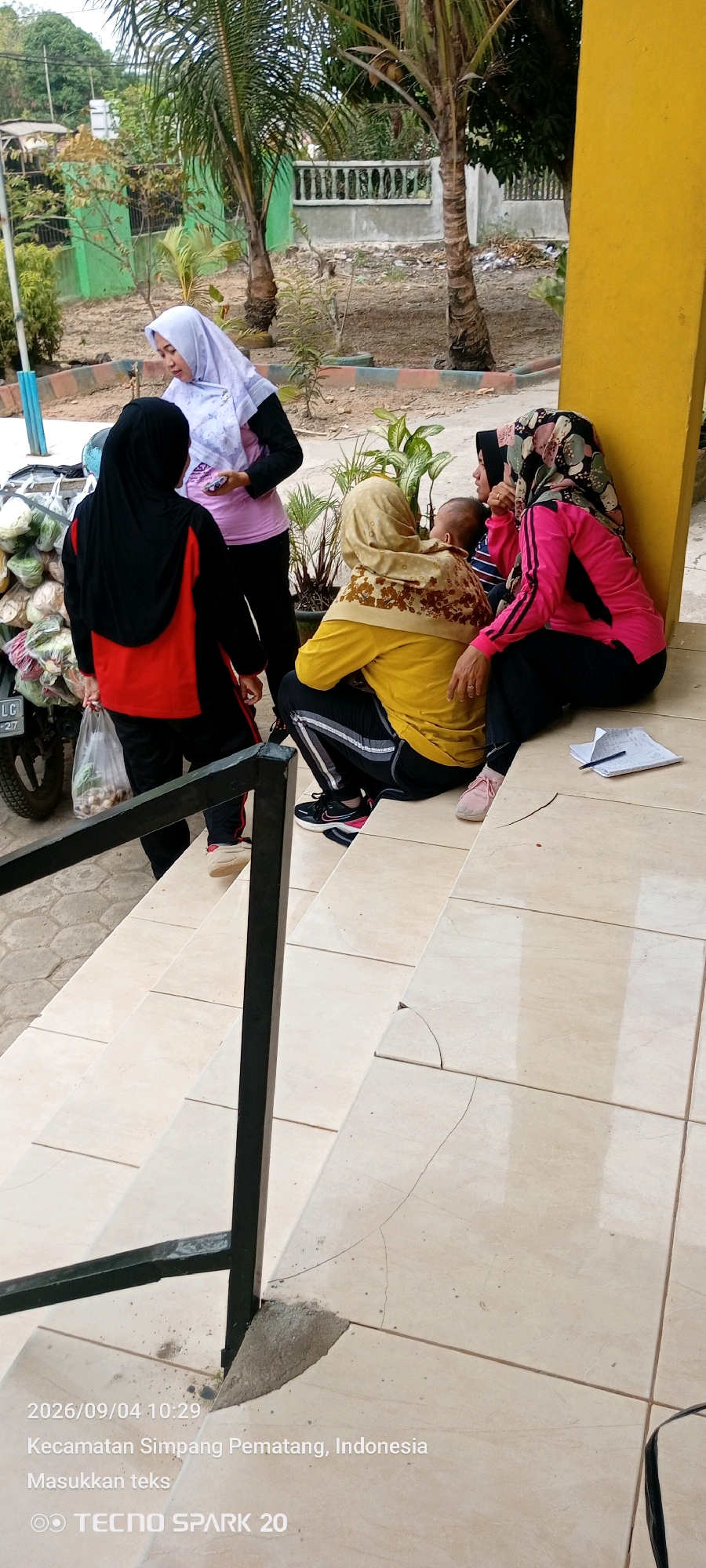 Four individuals are situated on the tiled steps of a building with a yellow exterior wall in Simpang Pematang, Indonesia. On the left, a person wearing a black hijab and a red and black jacket stands, holding a plastic bag filled with produce. To their right, another person in a light purple hijab is standing. In the center and right, two other individuals are seated on the steps, one holding a baby. The seated individuals are wearing headscarves, one in a tan hijab with a patterned overlay, and the other in a pink hijab with a floral pattern. The scene includes some greenery, a motorcycle loaded with vegetables, and a concrete structure. The lower portion of the image shows cracked tiles on the steps, with a metal railing in the foreground. A date stamp and location information indicating "2026/09/04 10:29, Kecamatan Simpang Pematang, Indonesia" are visible at the bottom left.