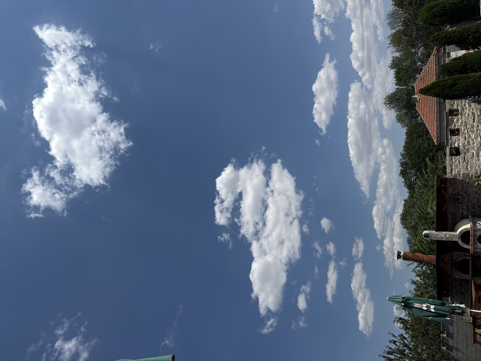 The image depicts a bright, sunny day in the city of Ekzarh Antimovo, Bulgaria. The sky is a clear, vibrant blue, dotted with fluffy white cumulus clouds. In the foreground, several structures are visible, suggesting an outdoor recreational or culinary space. A stone wall forms a prominent element, with a red-tiled roof building partially visible behind it. To the left, a green outdoor umbrella, branded with a logo, is partially deployed, casting a shadow. Adjacent to the umbrella is an outdoor oven structure, likely for pizza or bread, constructed from stone and brick, with a visible chimney. Further to the right, more stone construction and cypress trees are present. The background is filled with lush green trees, indicating a semi-rural or suburban setting within Ekzarh Antimovo. There are no people visible in the frame. The overall impression is one of a well-maintained outdoor area on a pleasant day.